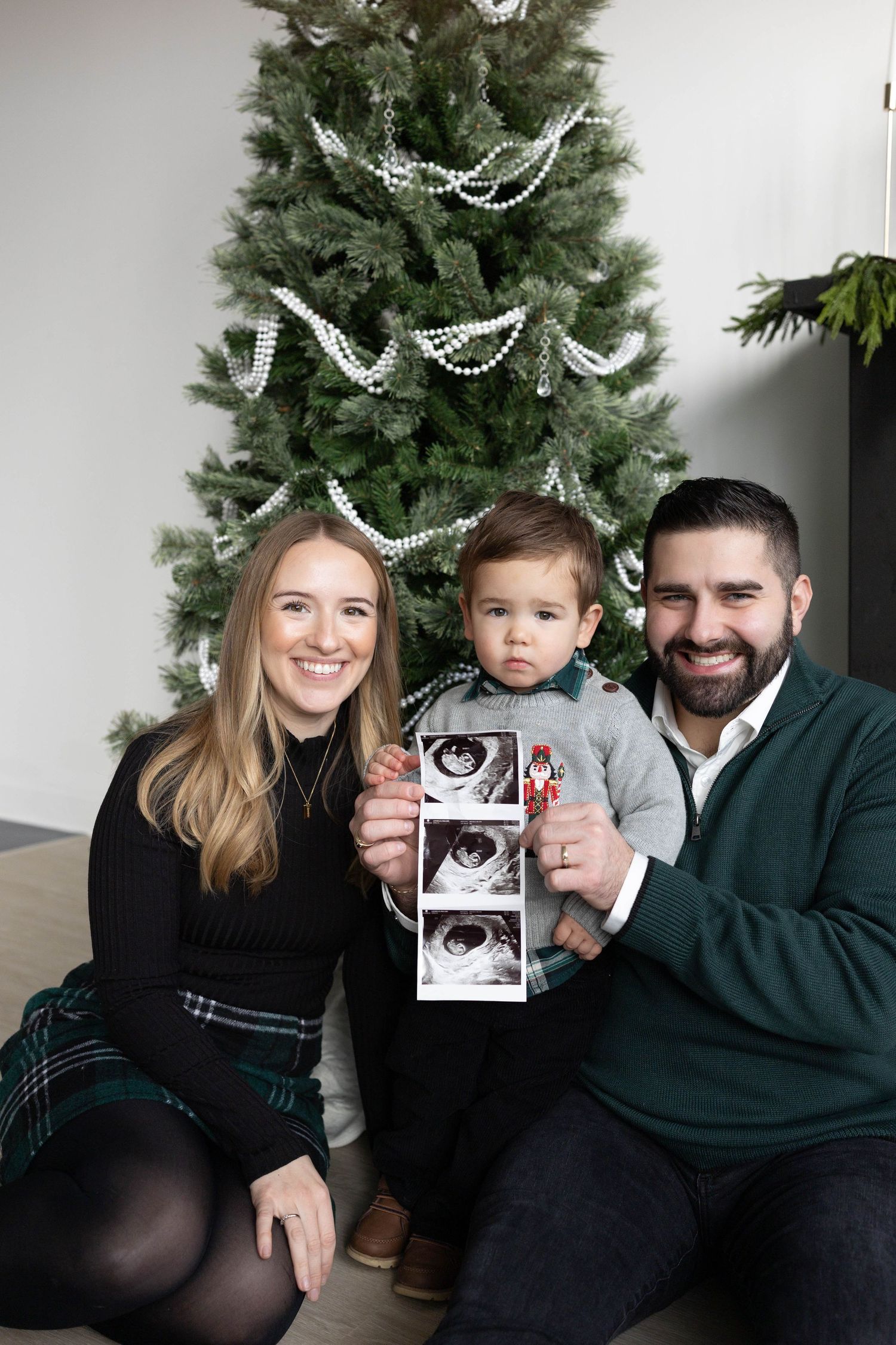 Family poses by Christmas tree with ultrasound photos while announcing pregnancy with festive silver garland decorations.