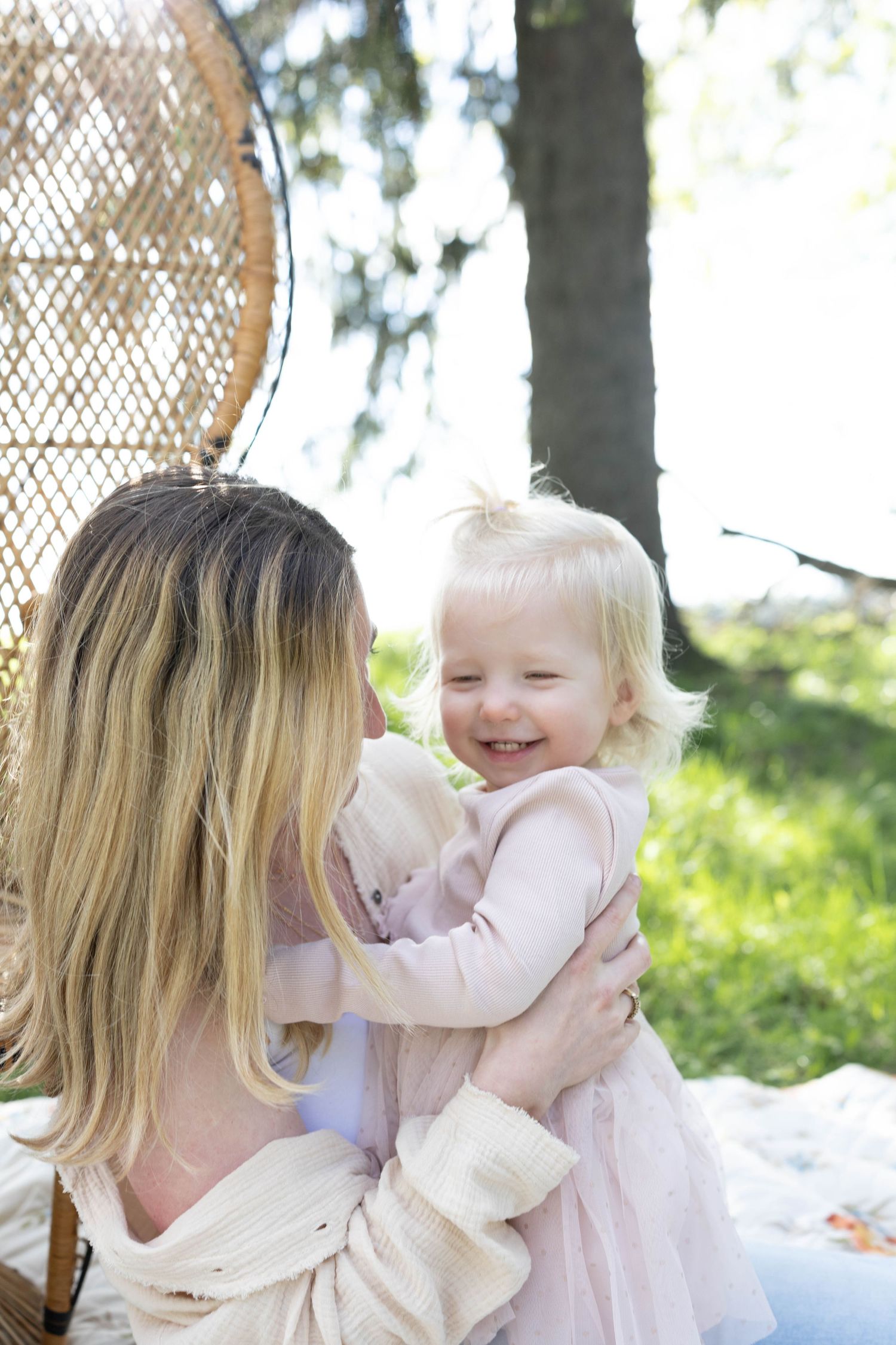 Tender outdoor moment between parent and delighted toddler in pink dress sitting on blanket with wicker chair.