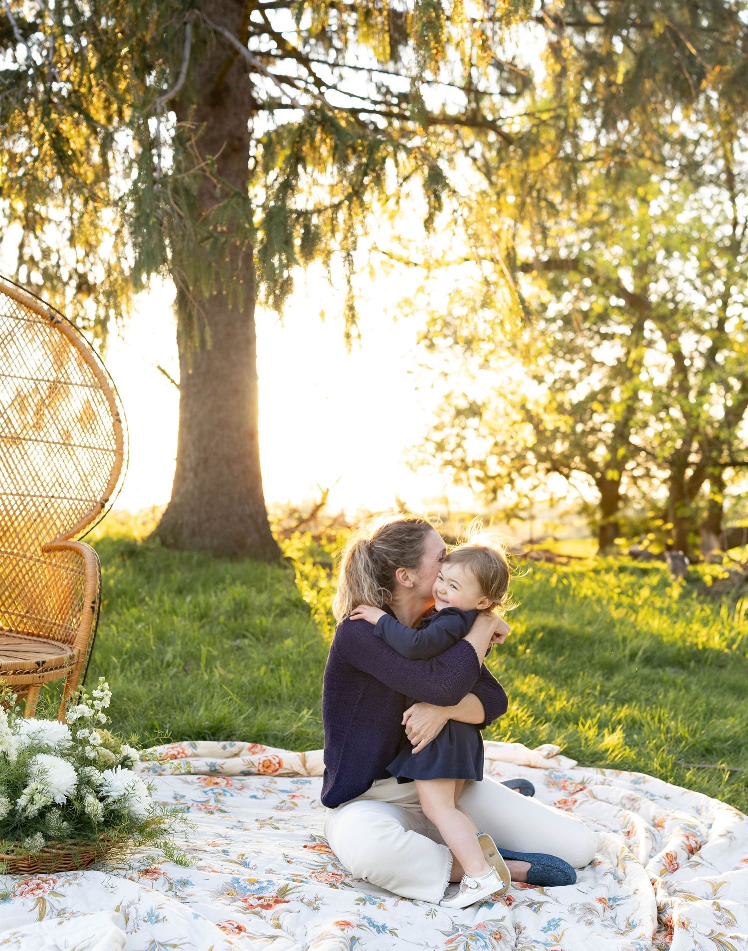 A peaceful picnic setup under a large tree at sunset with wicker furniture and cozy blankets spread on the grass.
