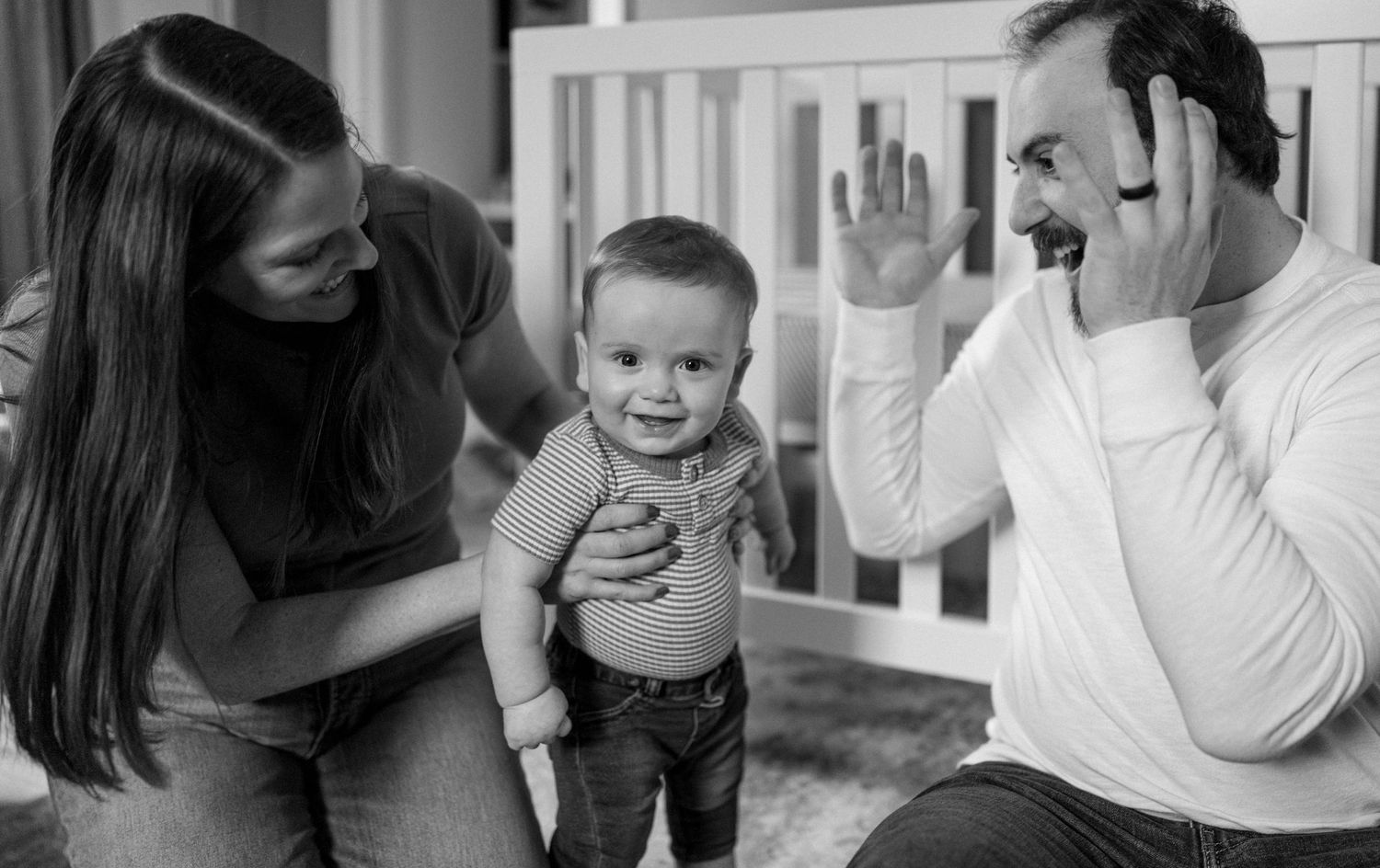 Black and white photo of parents interacting with their smiling baby who is standing and learning to balance.