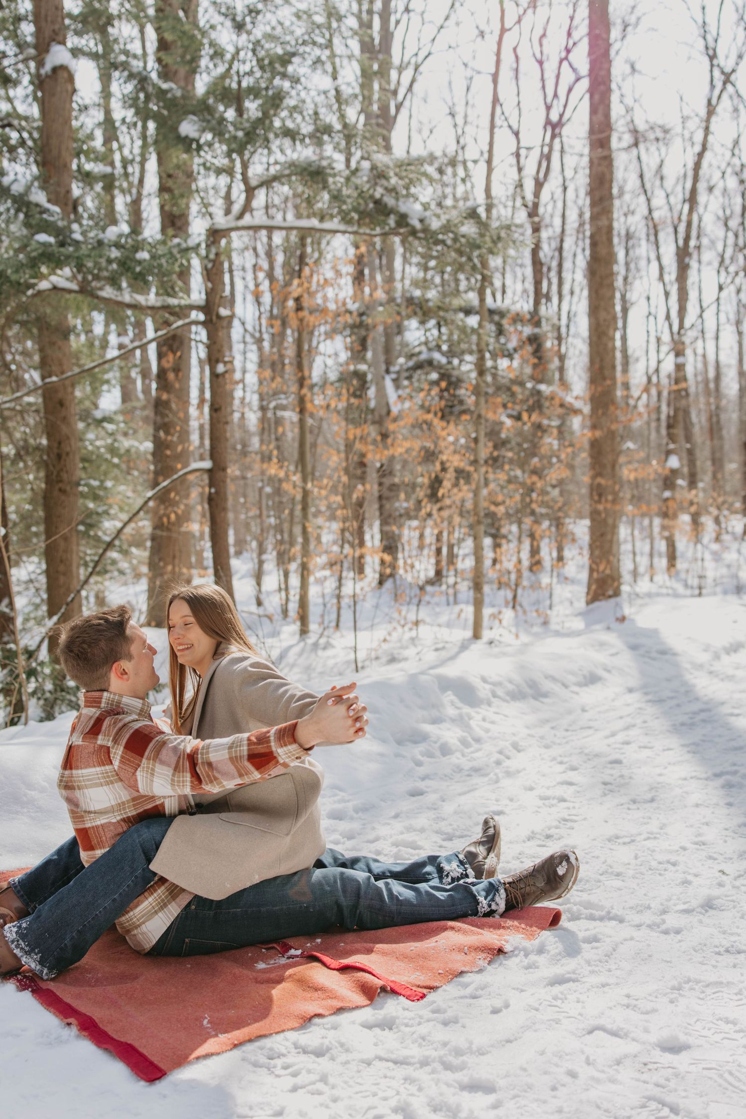 A couple relaxes on a red blanket in the snow-covered forest during winter while enjoying a romantic moment together.