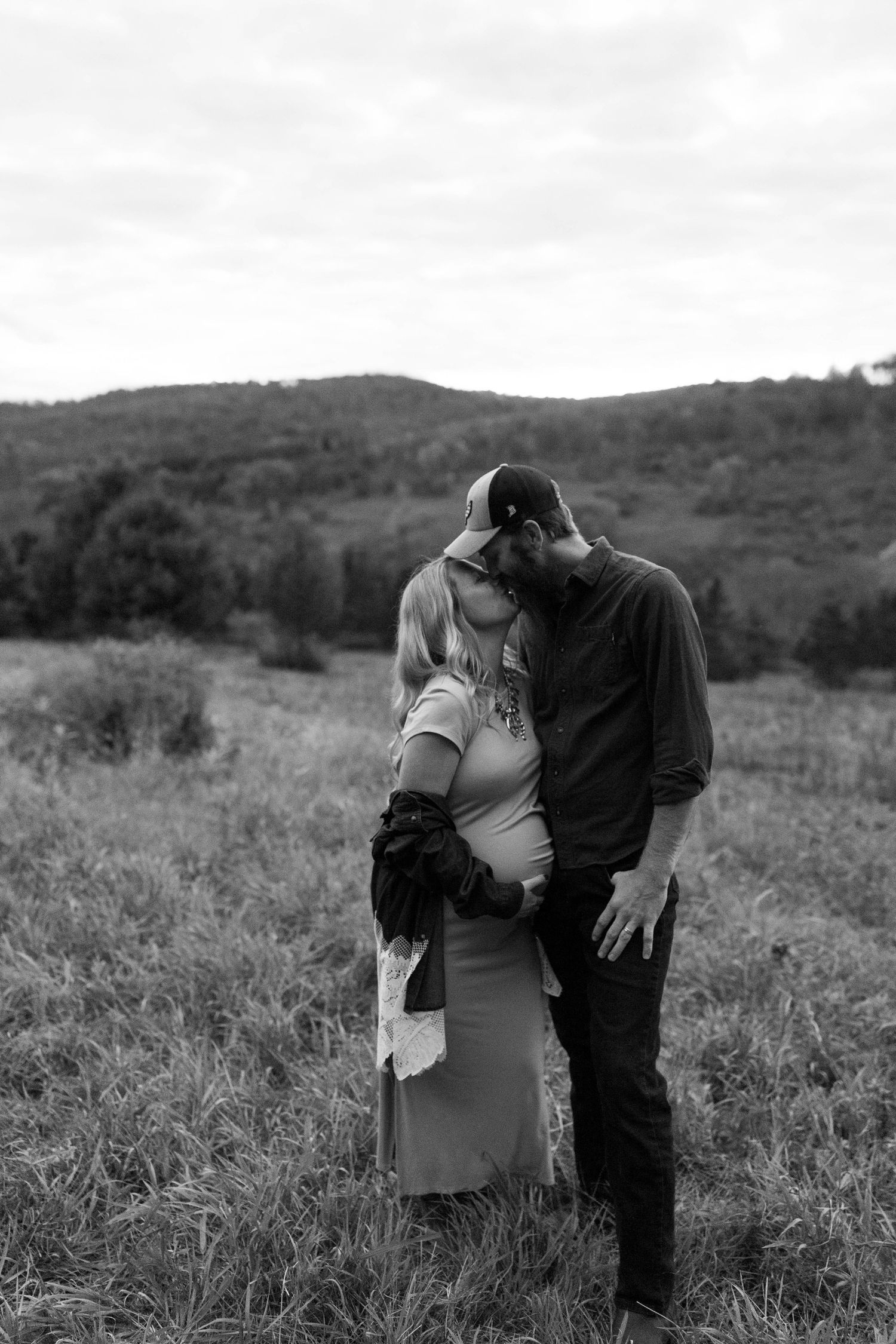 Romantic couple embraces in a scenic field with rolling hills in the background during an autumn photoshoot.