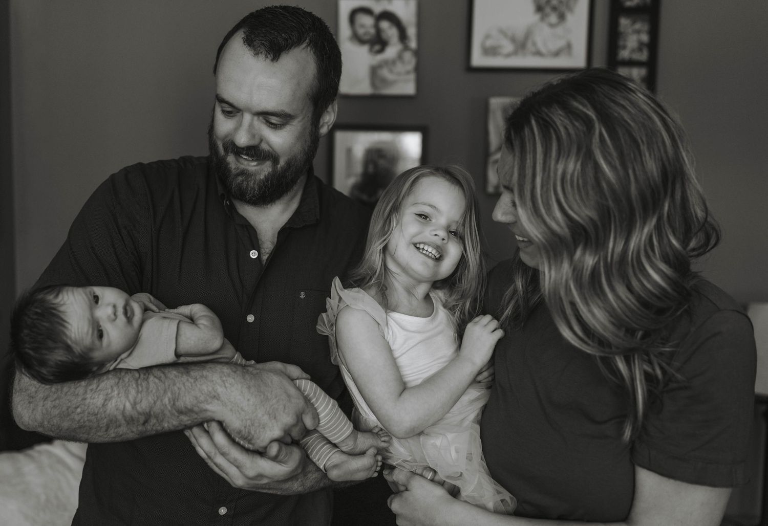 Black and white family photo of parents smiling with newborn baby and young daughter in a cozy home setting.