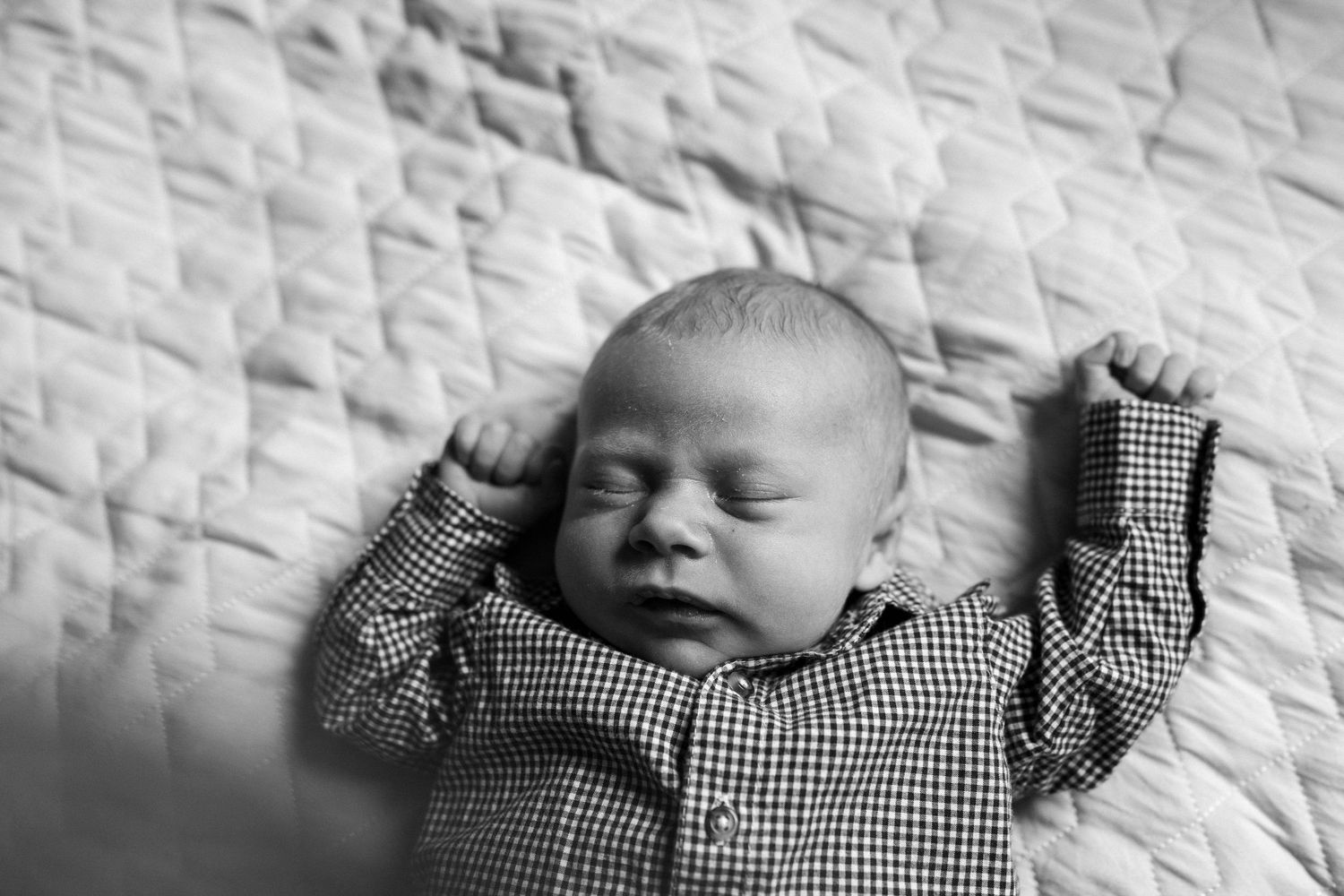 Black and white photo of a peaceful sleeping newborn baby in a checkered shirt lying on a quilted blanket.