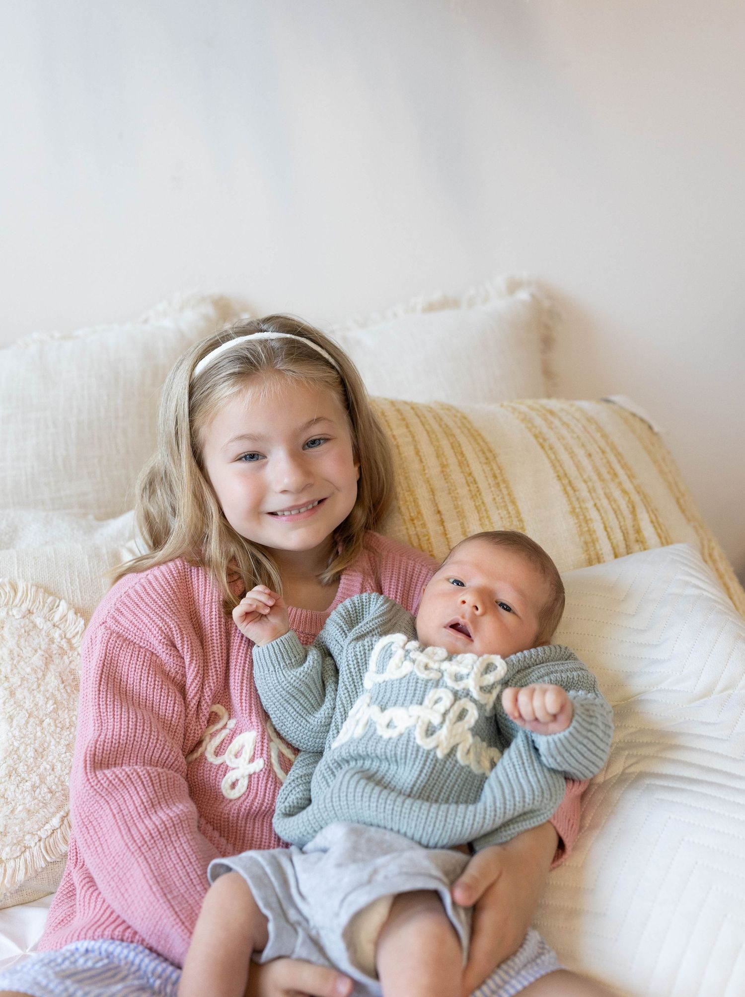 Two children in cozy sweaters cuddle together on a cream-colored couch, showing a sweet moment of affection.