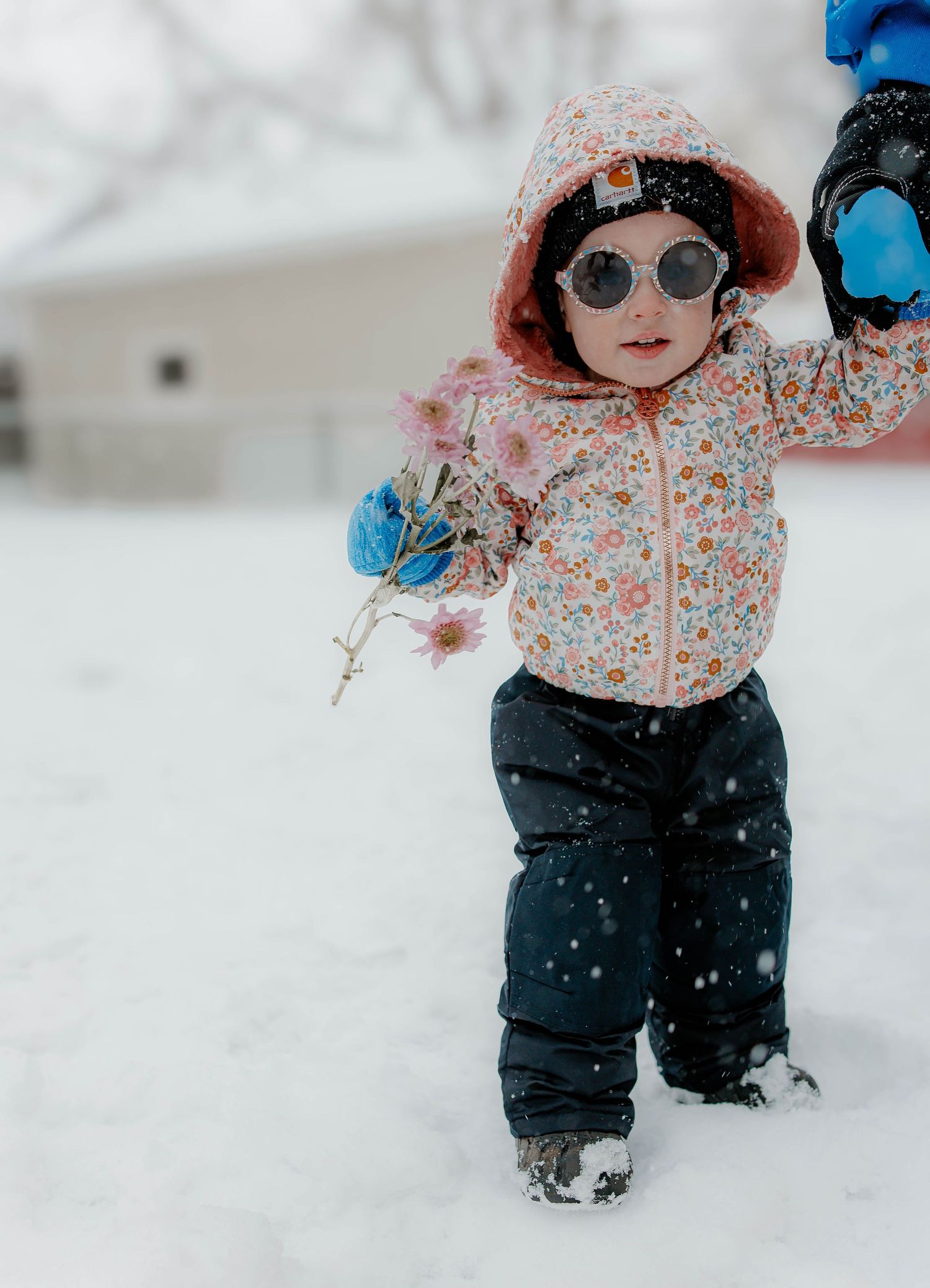 Small child in floral print jacket and snow pants plays outside in winter with sunglasses and mittens.