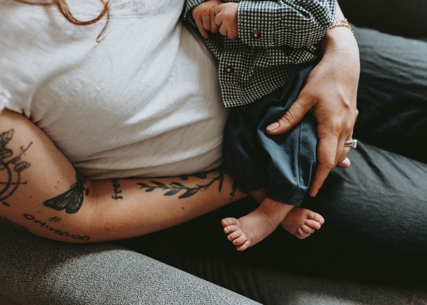 Tattooed mother holding newborn baby with tiny bare feet on a gray couch.