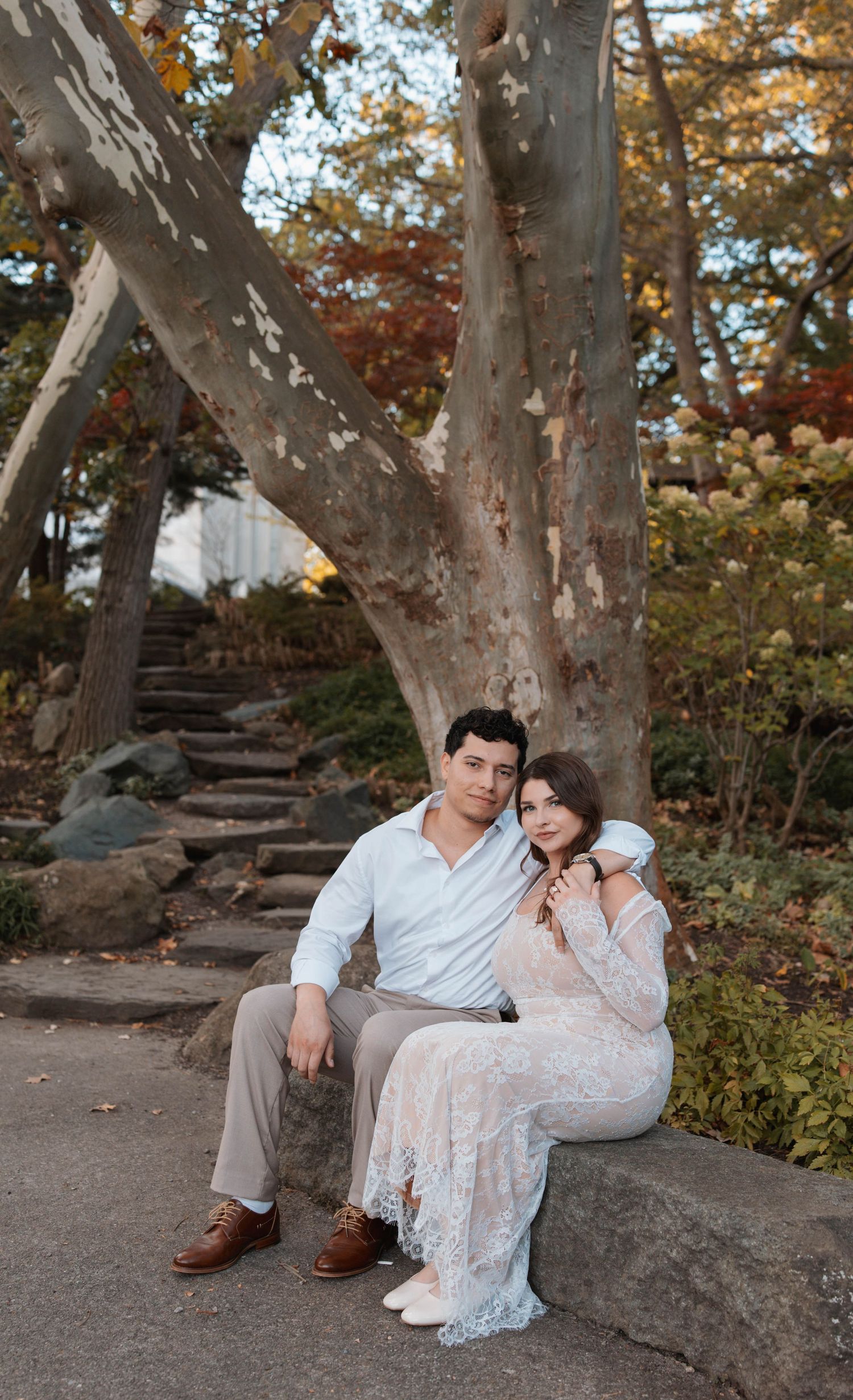 A romantic couple sits on stone steps surrounded by autumn trees in a park setting during a photoshoot.