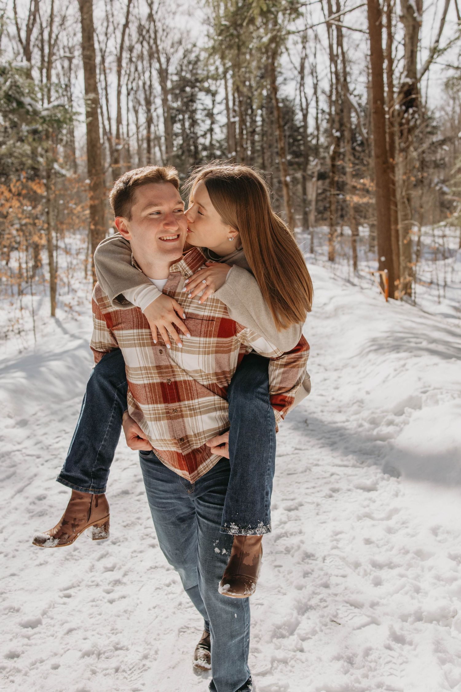 Romantic couple embracing and playing in a snowy forest trail during winter wearing casual plaid and denim.
