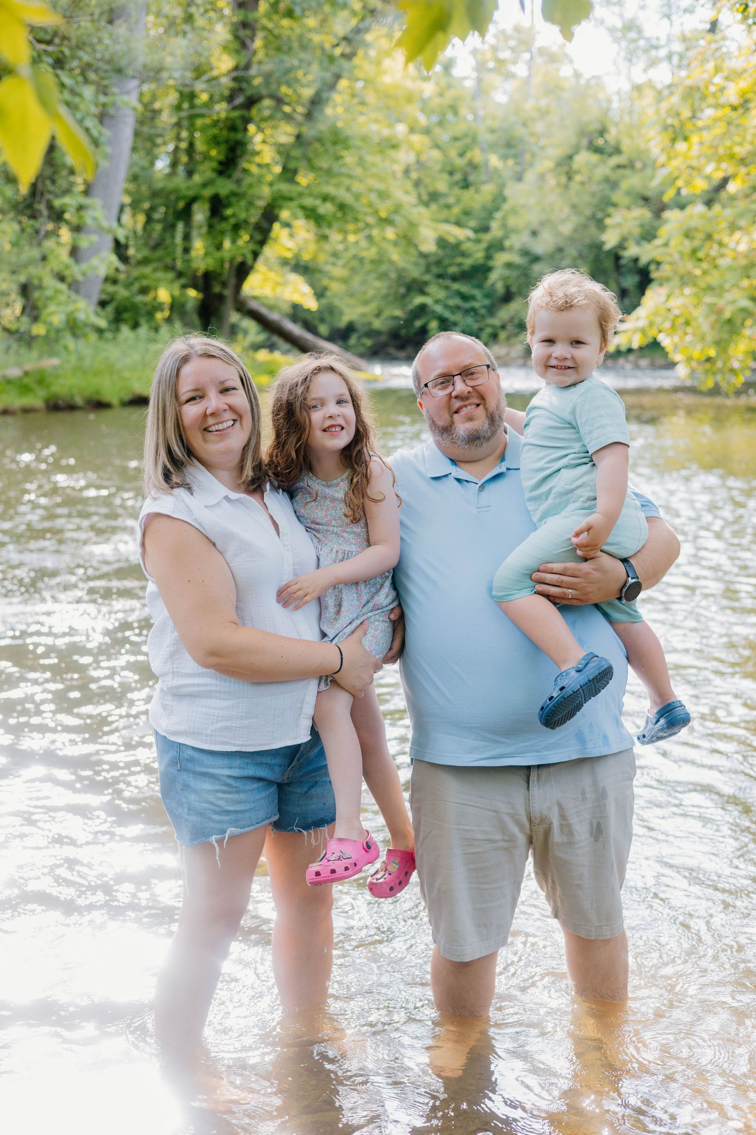 Happy family of four standing in a shallow river surrounded by lush green trees on a sunny day.