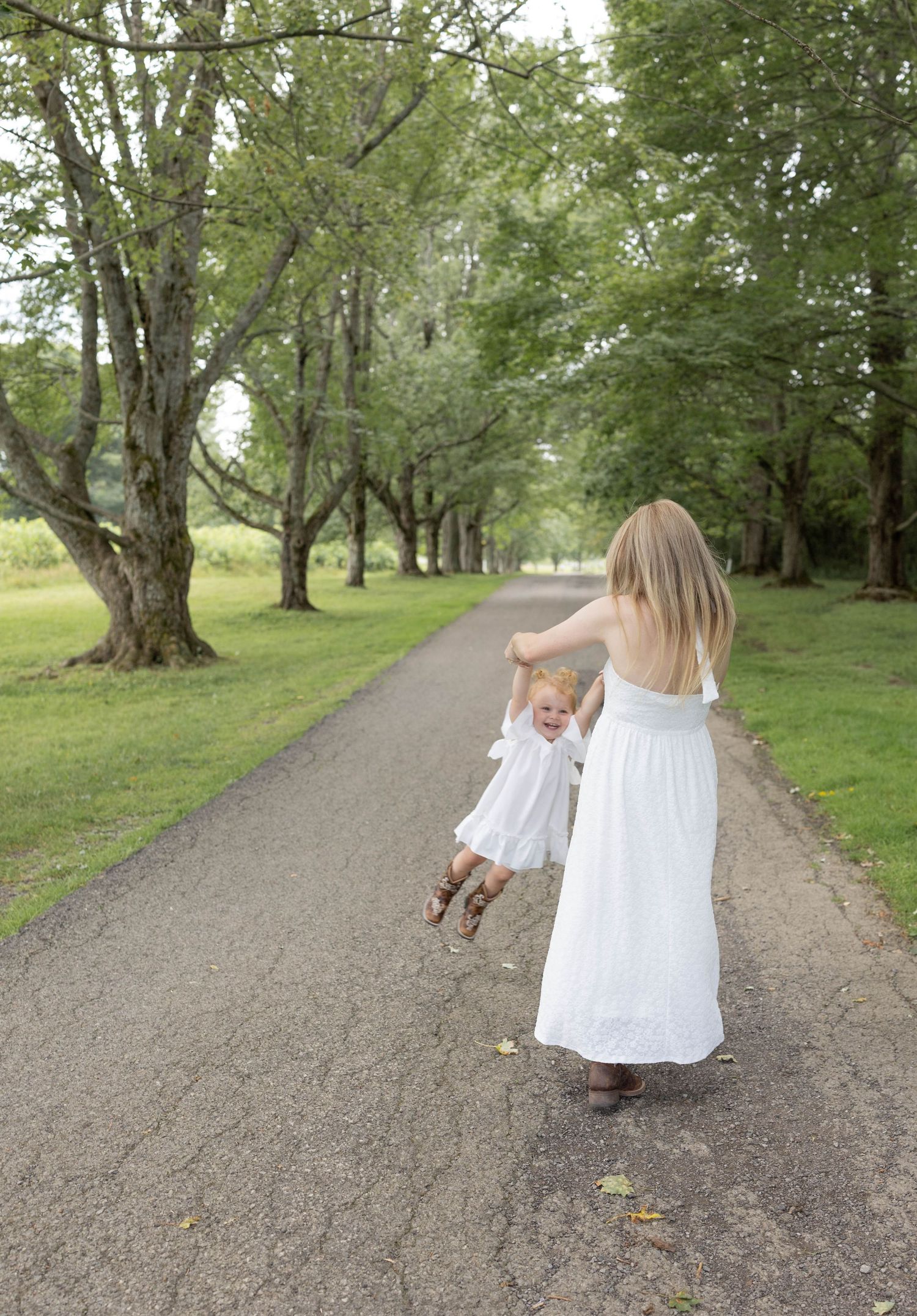 A mother in a white dress swings her toddler on a tree-lined path outdoors.