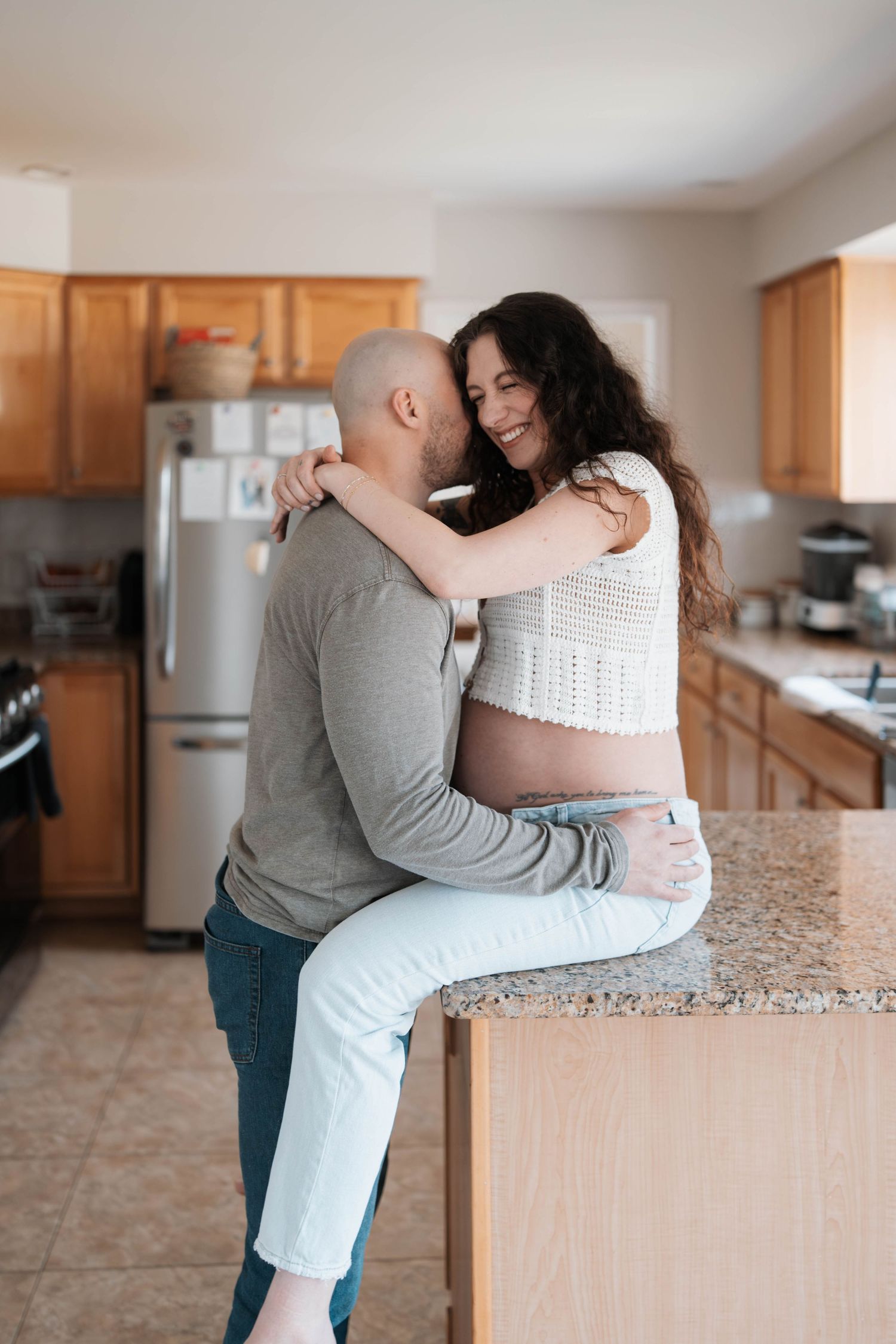 A tender moment between a couple embracing in their kitchen as she sits on the granite counter.