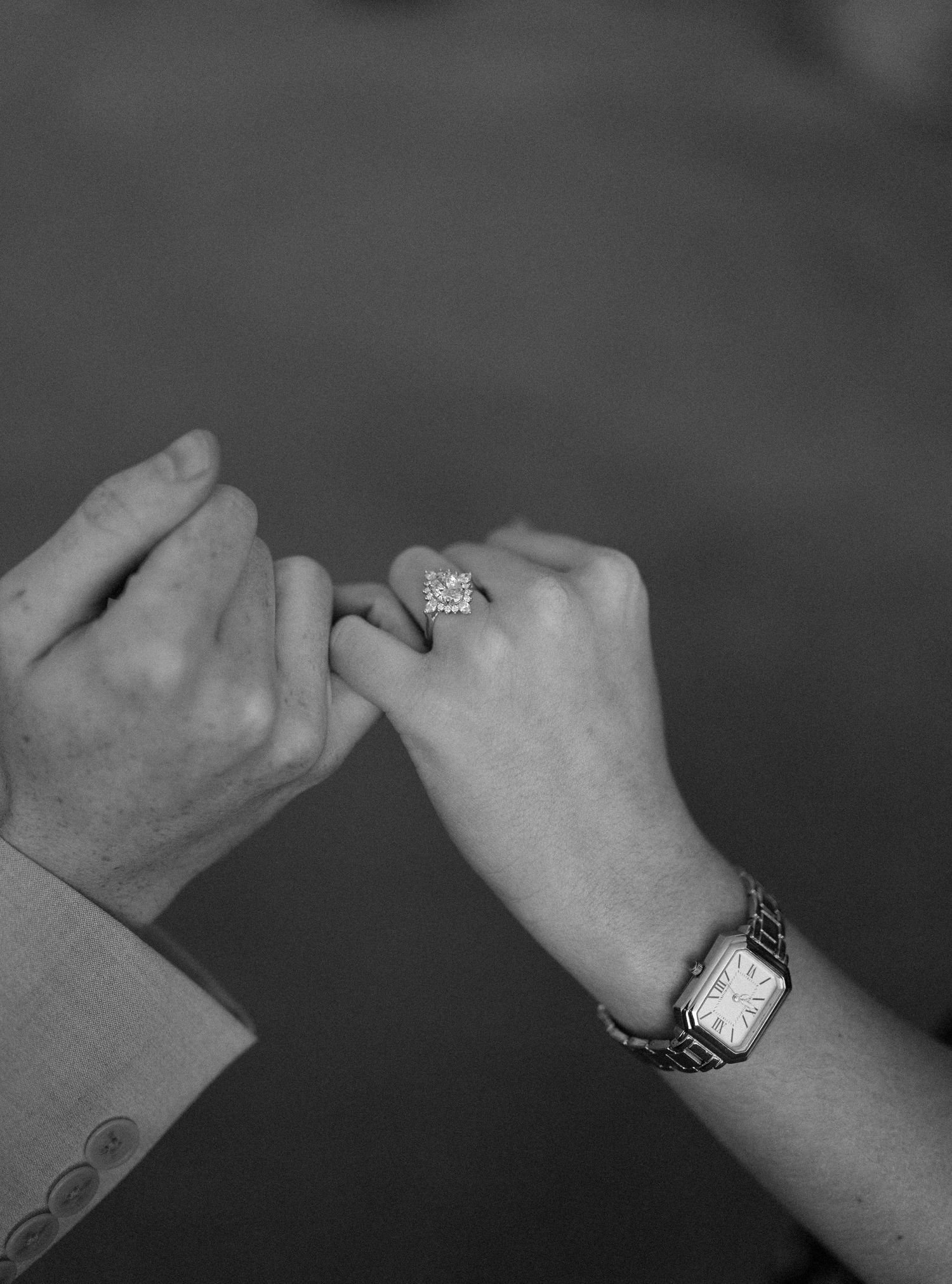 Two clasped hands joining pinky fingers in a close-up black and white shot, with one hand wearing a ring and watch.