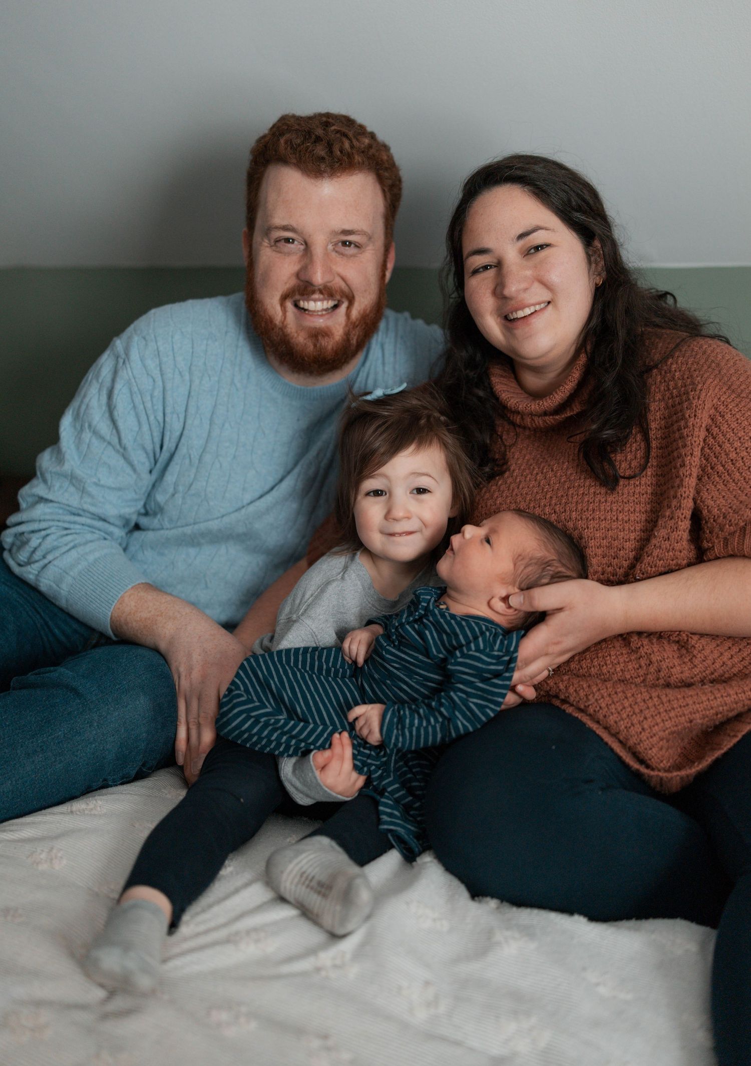 A family of four sitting together on a bed wearing warm casual sweaters in a cozy relaxed pose.
