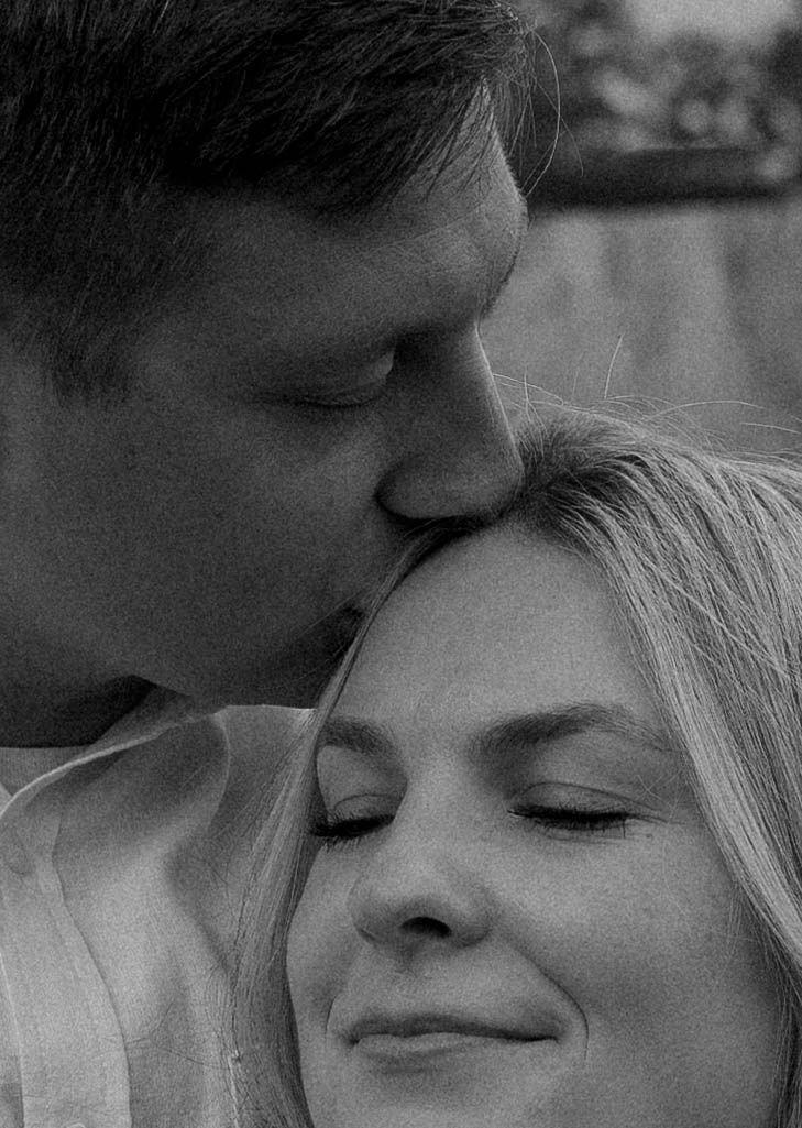 Black and white photo of a man tenderly kissing a smiling woman on the forehead in a romantic outdoor setting.