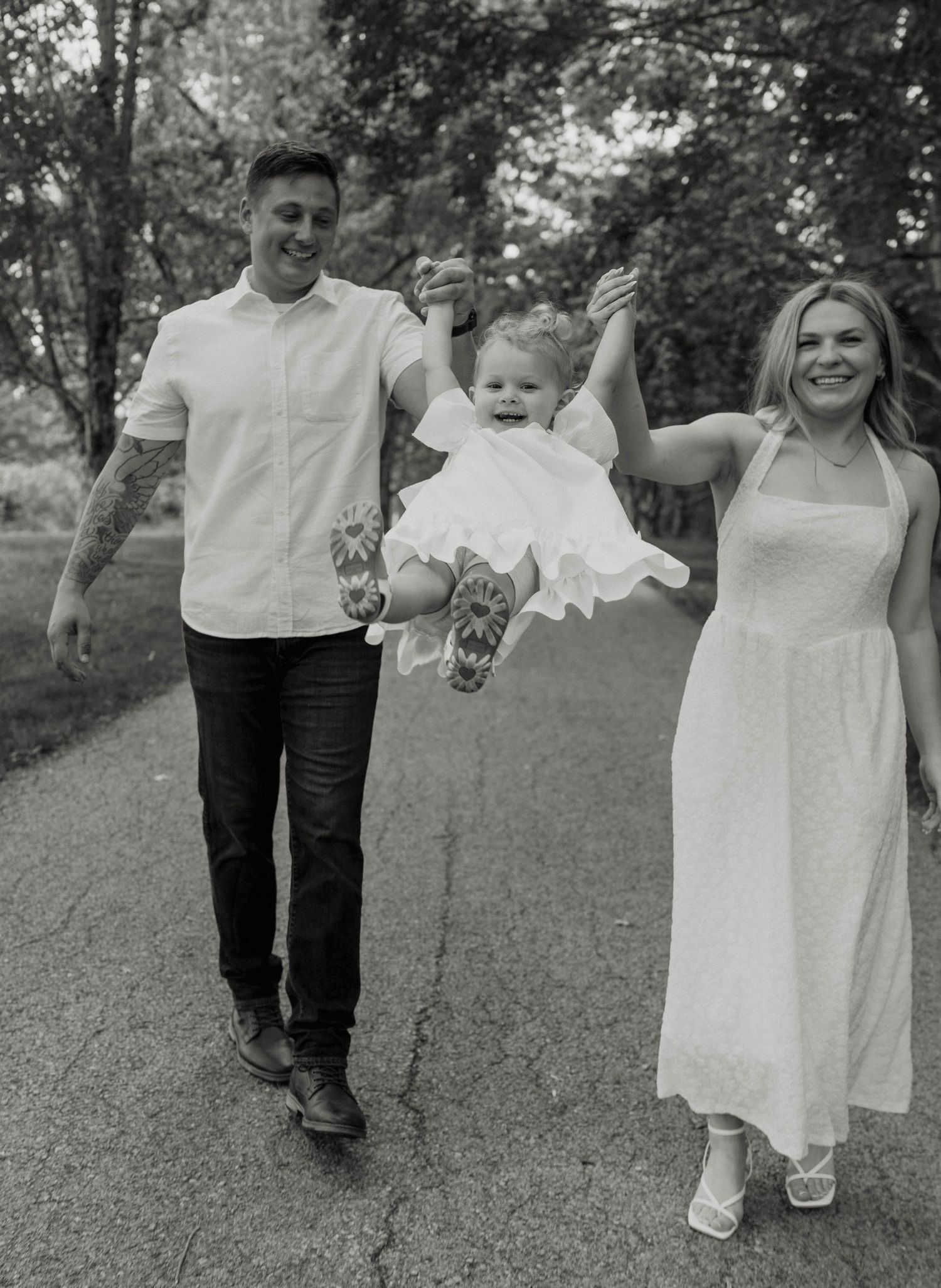 Black and white photo of a happy couple swinging their toddler by the hands on a tree-lined path.