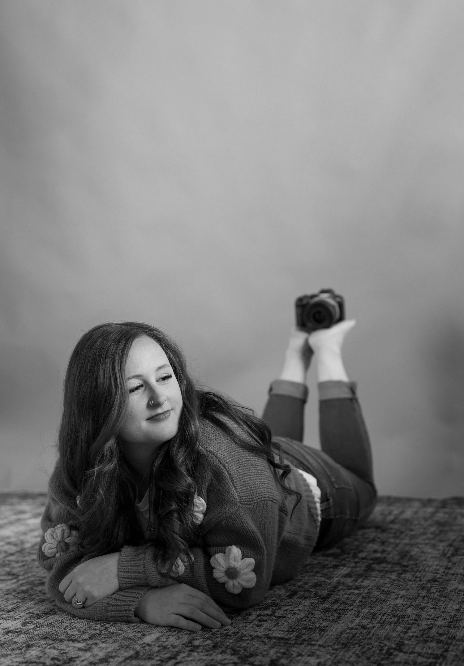 Black and white photo of a young woman lying on the floor holding a camera, wearing a floral sweater.