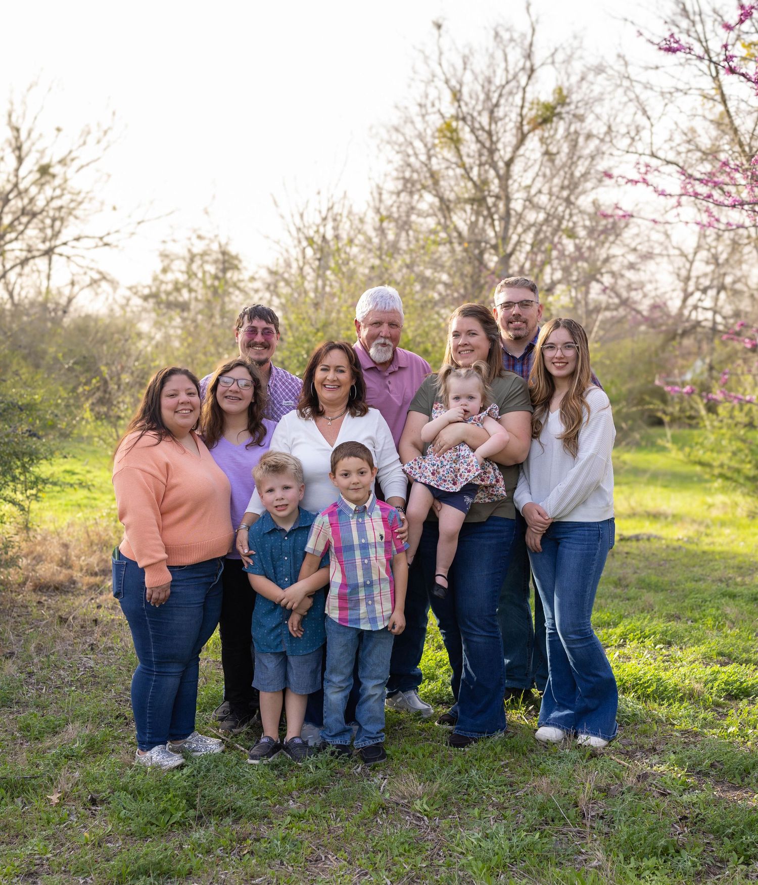 A multi-generational family stands together outdoors in springtime with blossoming trees and green grass in the background.