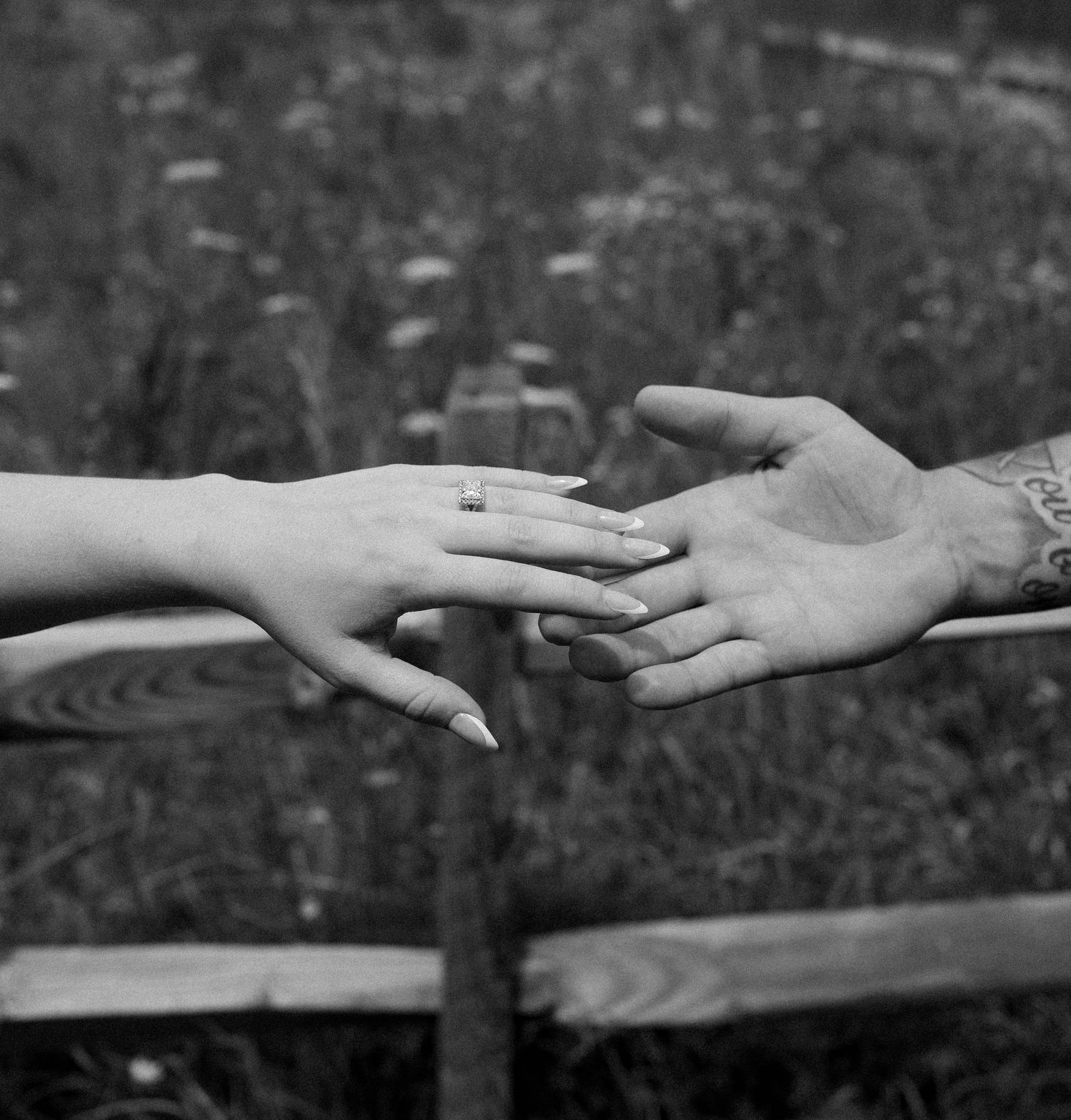 Black and white photo of two hands gently touching, one wearing a ring, with a tattooed wrist visible.