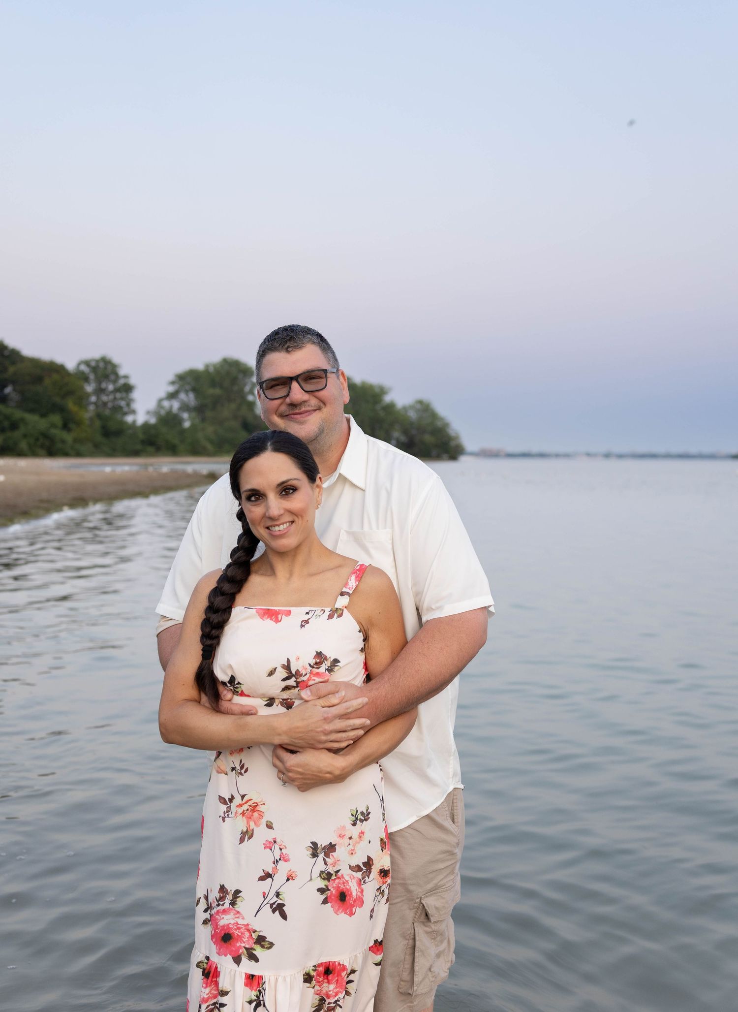 Couple embracing and smiling on a serene beach at dusk, woman in floral dress with braid, man in white shirt.