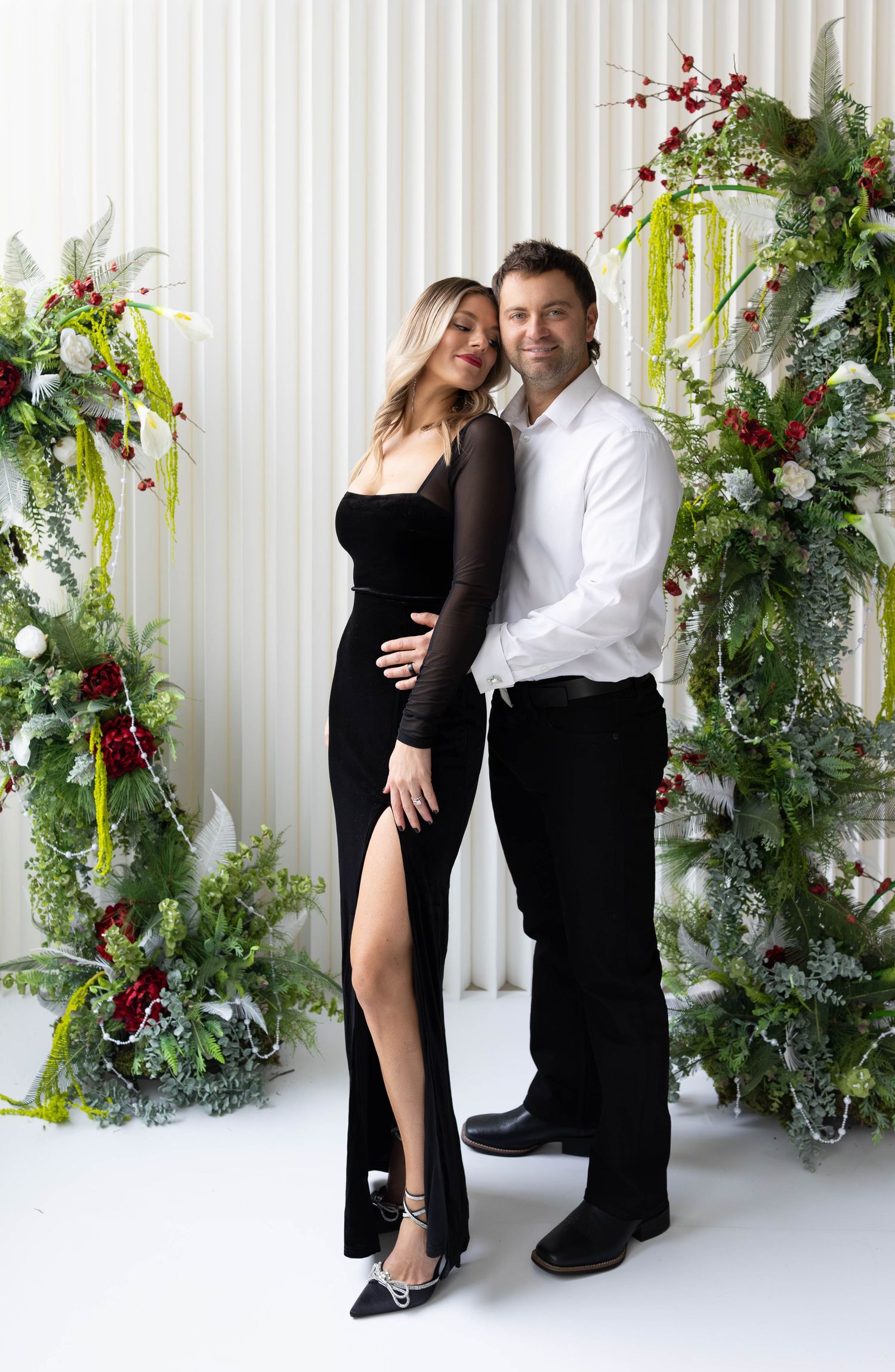Romantic couple poses in elegant black and white outfits against a white wall decorated with holiday greenery and red berries.