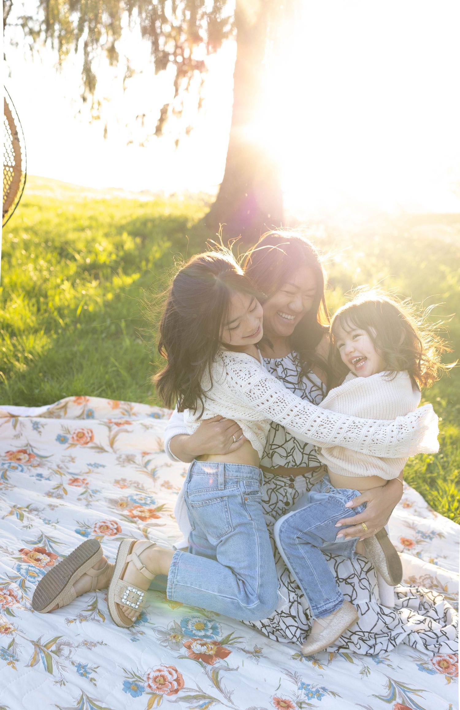 Three people share joyful moments together on a floral blanket in a sunlit grassy field during golden hour.