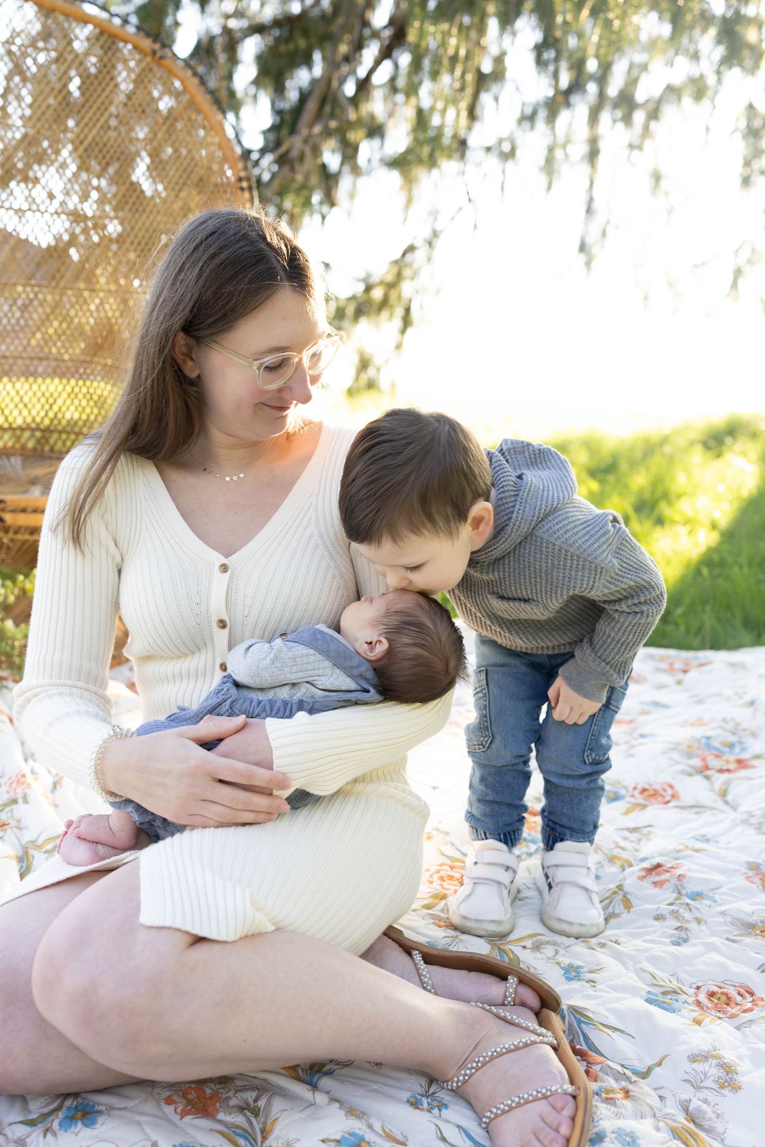 A mother sits outdoors on a blanket with two children on a sunny day under a willow tree.
