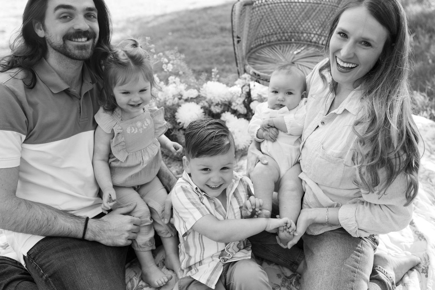 Black and white outdoor family portrait showing casual group sitting together on wicker chair in natural setting.