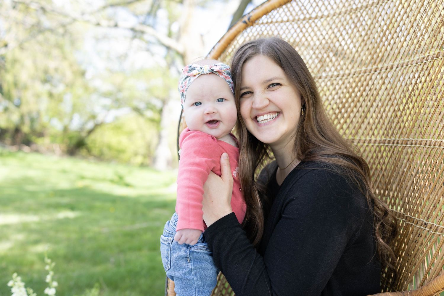 Mother and baby pose outdoors against wicker backdrop in sunny garden setting.