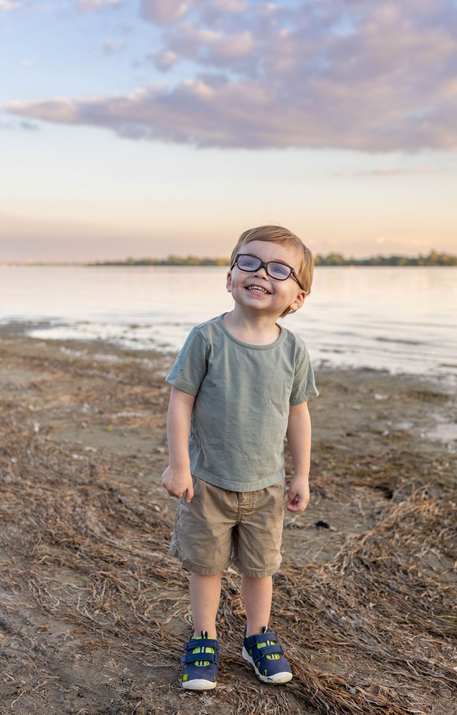 A toddler in casual clothes enjoys a beach sunset while looking up at the sky with wonder.