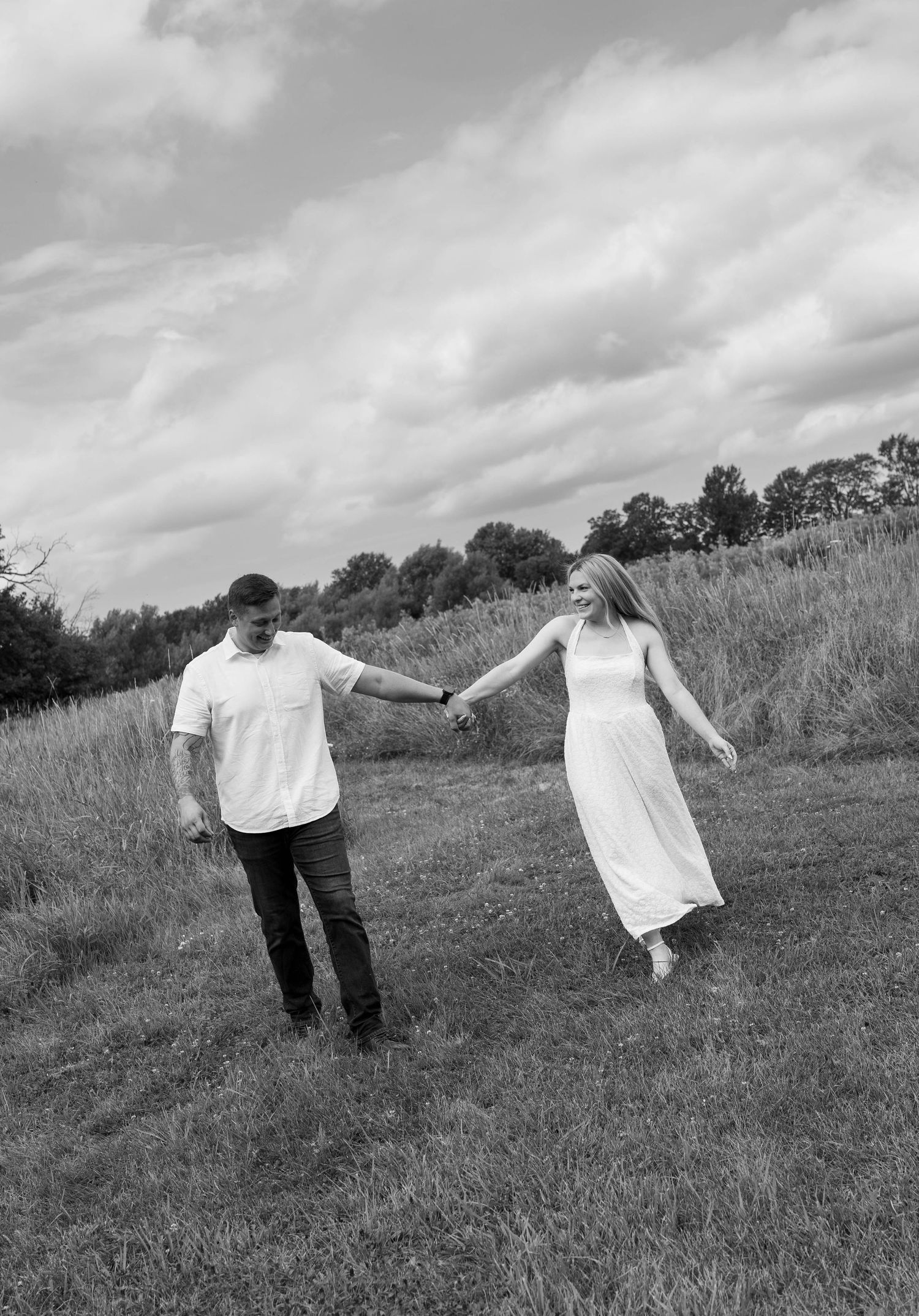 Black and white photo of a couple holding hands and walking through a grassy field under a cloudy sky.