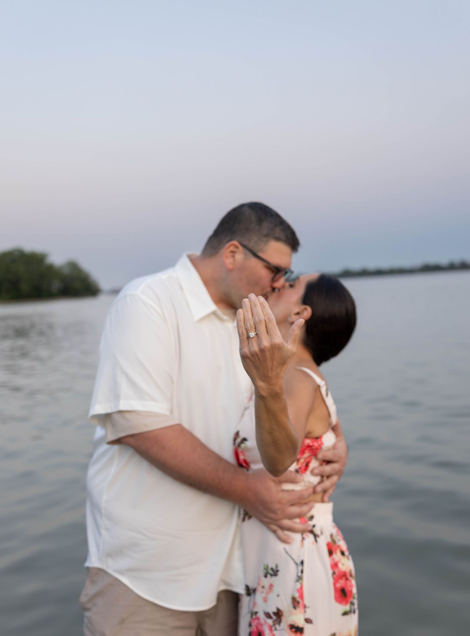 Couple sharing a romantic kiss by the lake, woman showing engagement ring, man in white shirt embracing her.