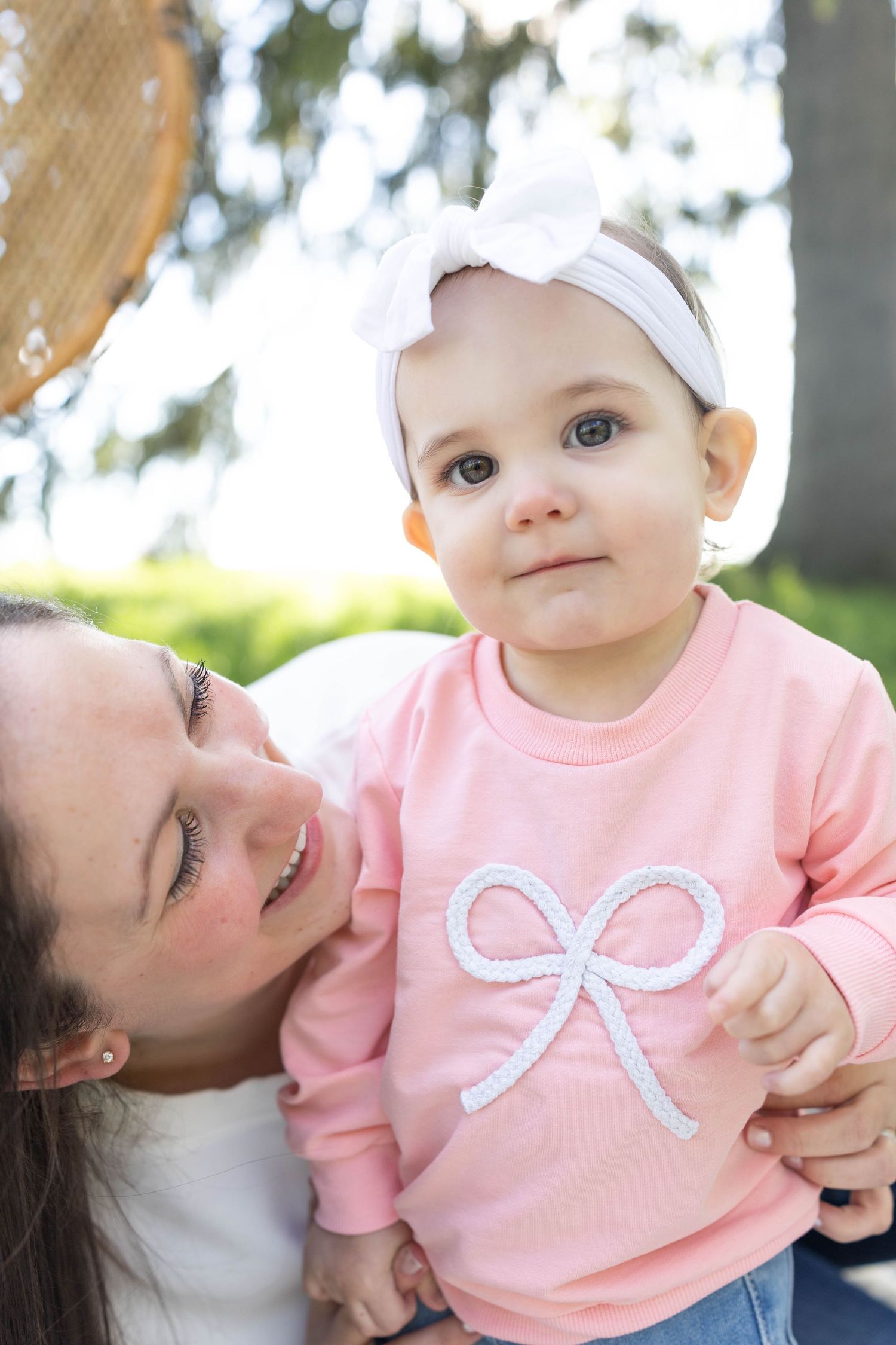 A baby wearing a pink sweatshirt with bow design and white headband outdoors on a sunny day.