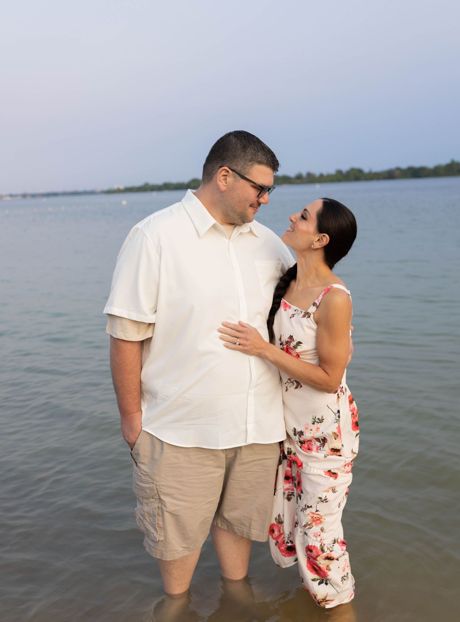 Couple standing in shallow water at a lakeside, smiling at each other, man in white shirt and woman in floral jumpsuit.