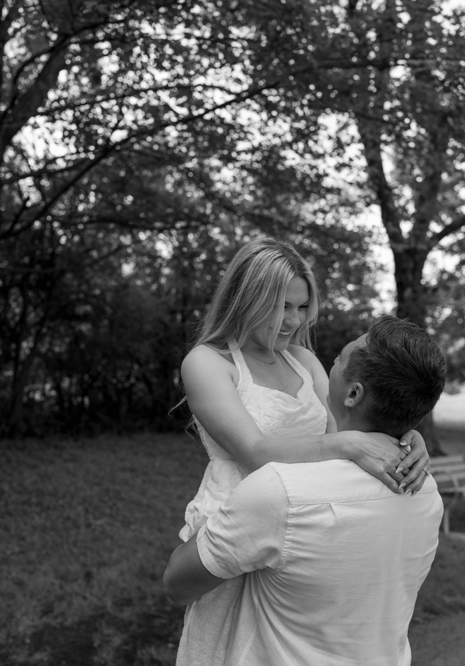 Black and white photo of a man lifting a woman in a white dress in an outdoor park setting.