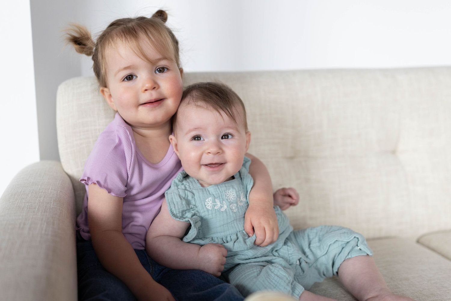 Two young sisters sitting on a cream sofa, older toddler in purple shirt hugging smiling baby in teal romper.