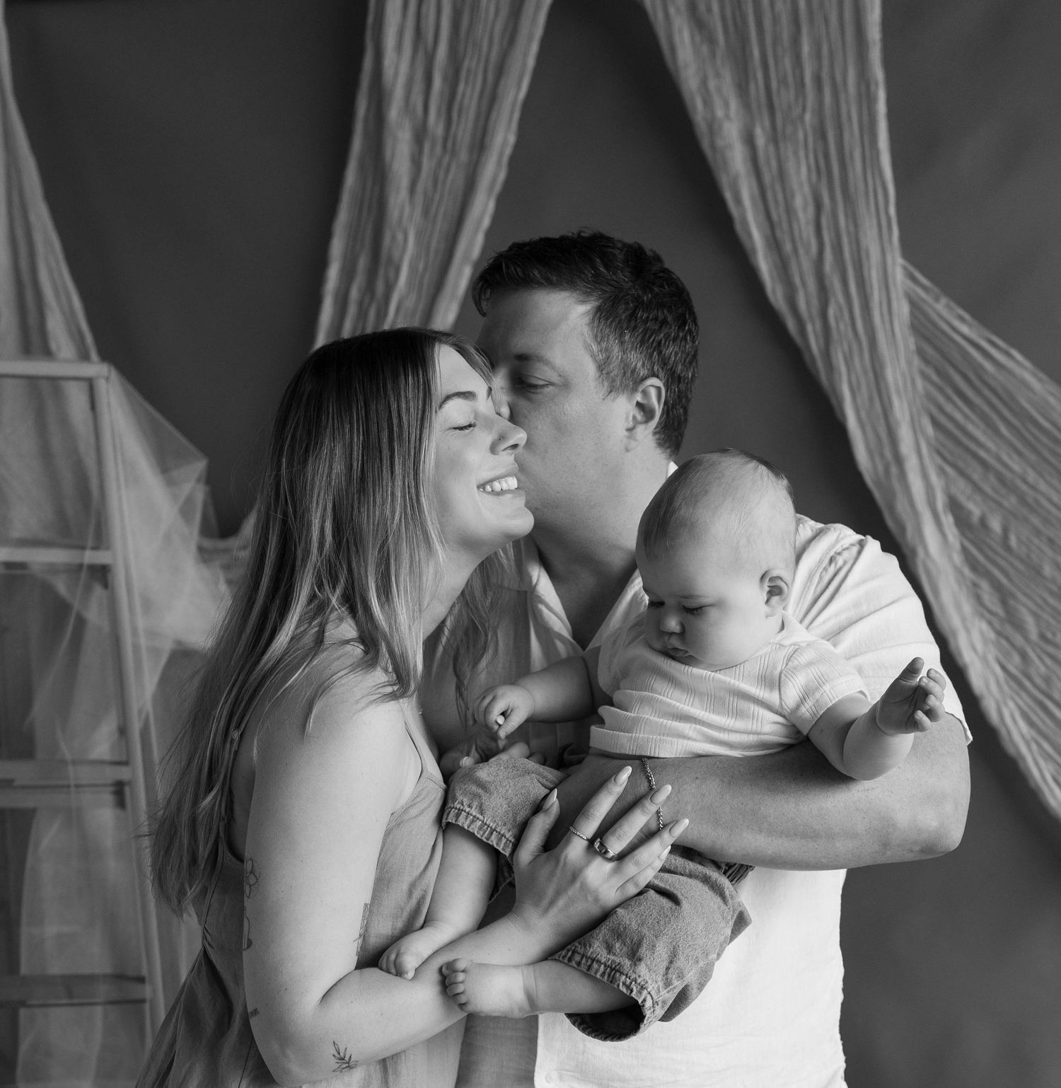 Black and white photo of a couple sharing a tender kiss while holding their newborn baby.
