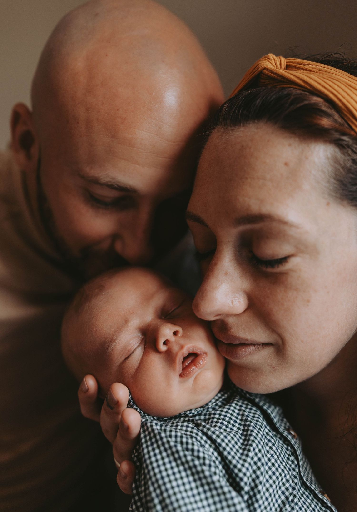 New parents tenderly kissing their sleeping newborn baby dressed in a gingham outfit.