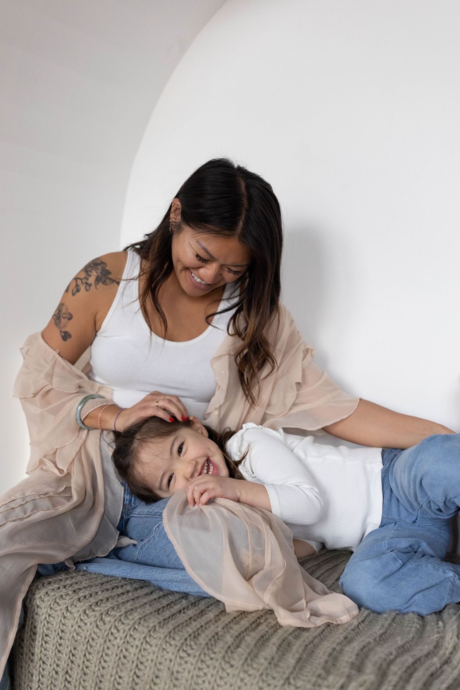 Two women laughing and relaxing together on a bed, one lying down while the other sits beside her smiling.