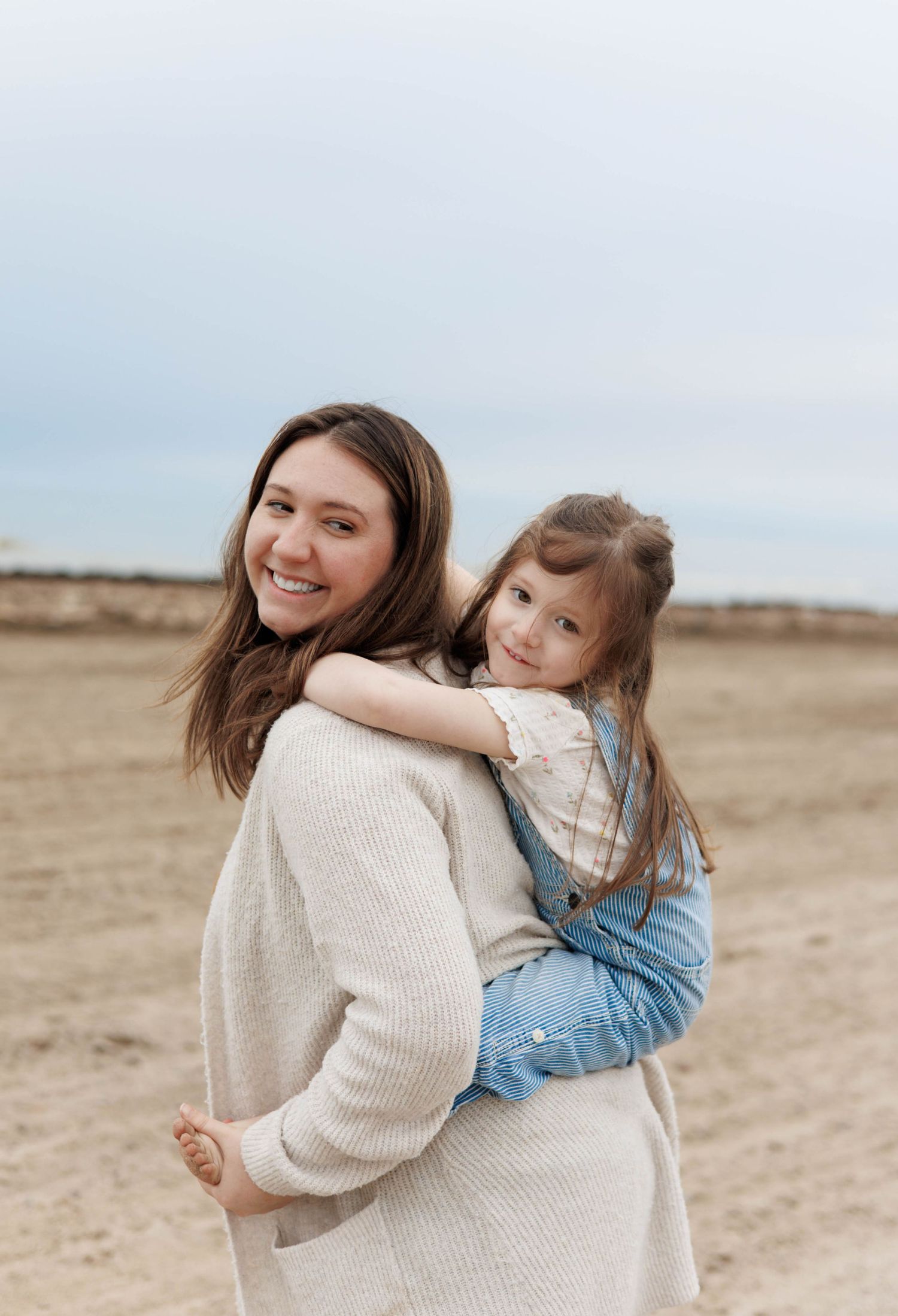A smiling mother holds her young daughter on a sandy beach, both wearing casual outfits under a cloudy sky.