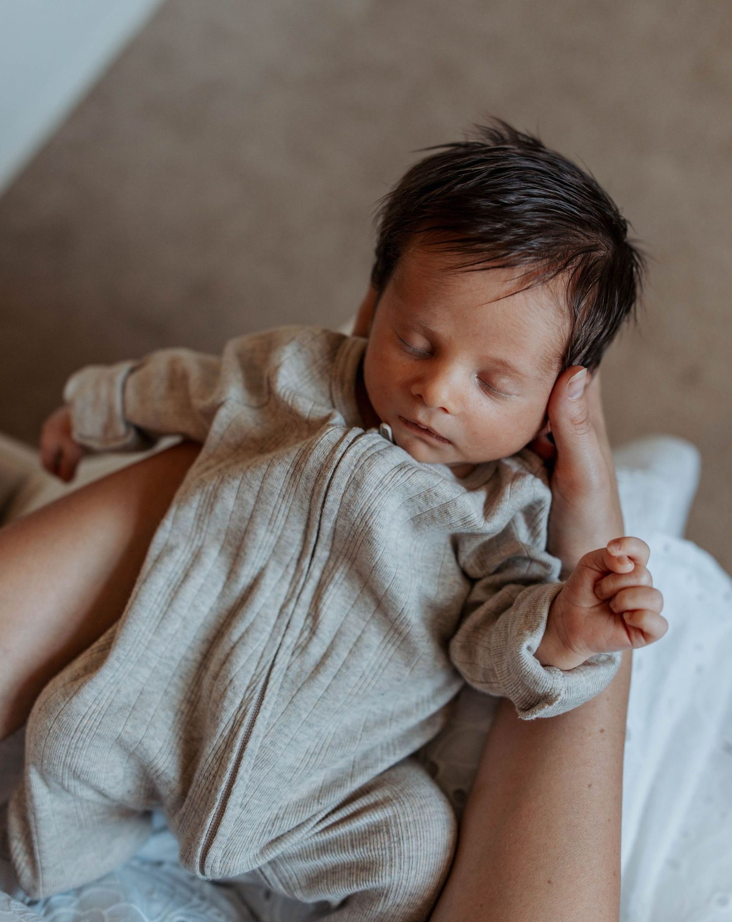 Sleeping newborn baby in a beige knit romper resting peacefully on a white blanket.