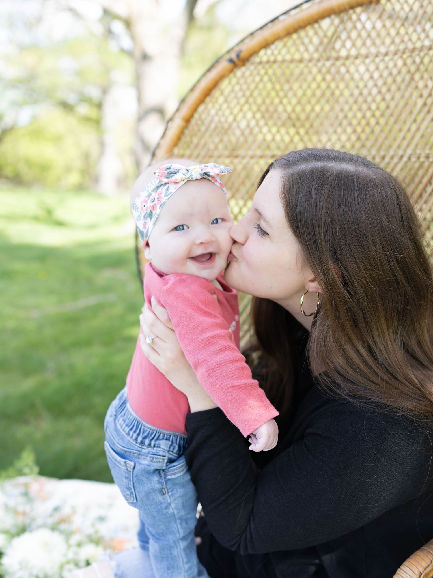 A tender moment captured outdoors as baby in coral sweater shares a joyful smile while being held against a wicker chair.