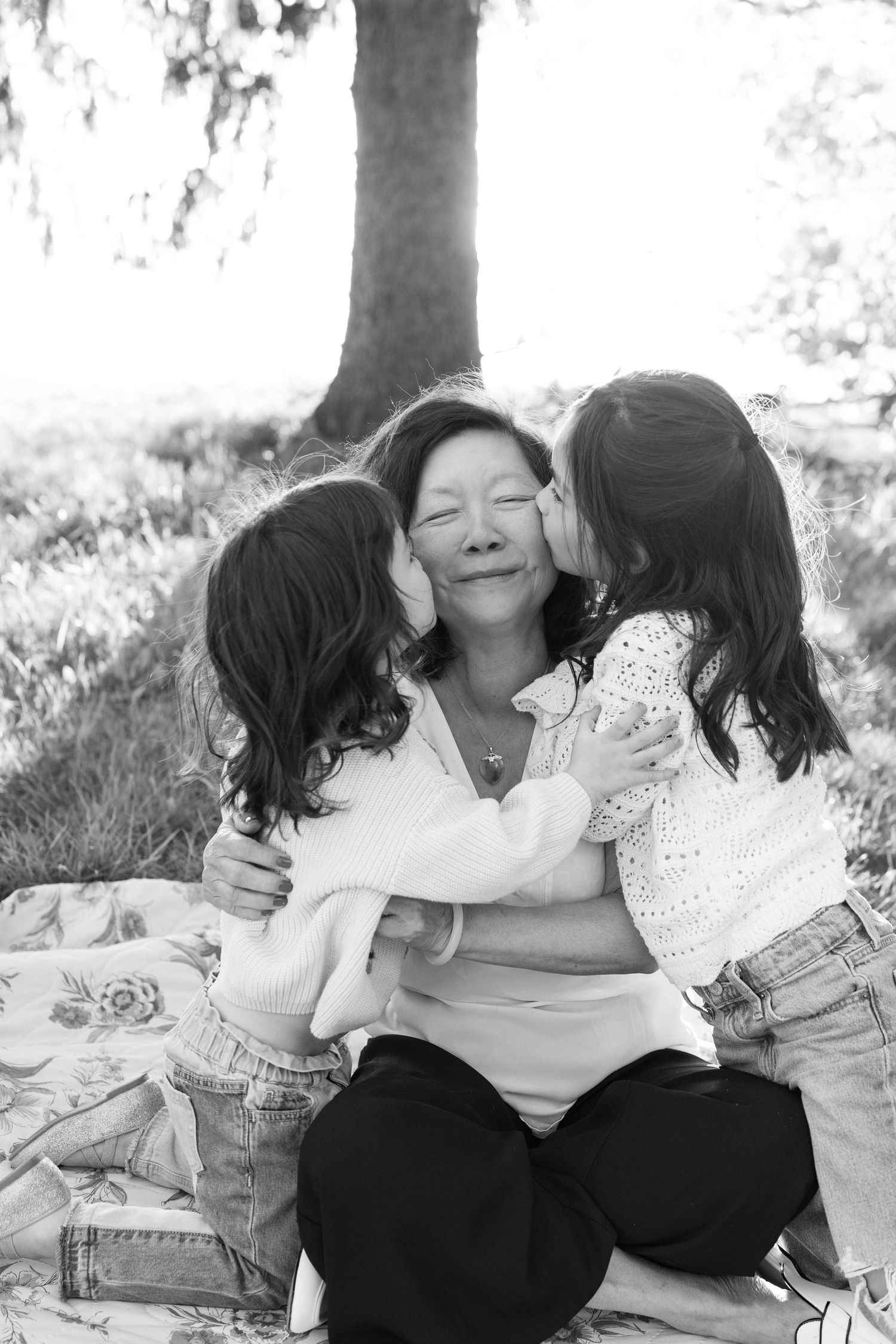 Two children affectionately hugging loved one while sitting together on a blanket outdoors in black and white photography.