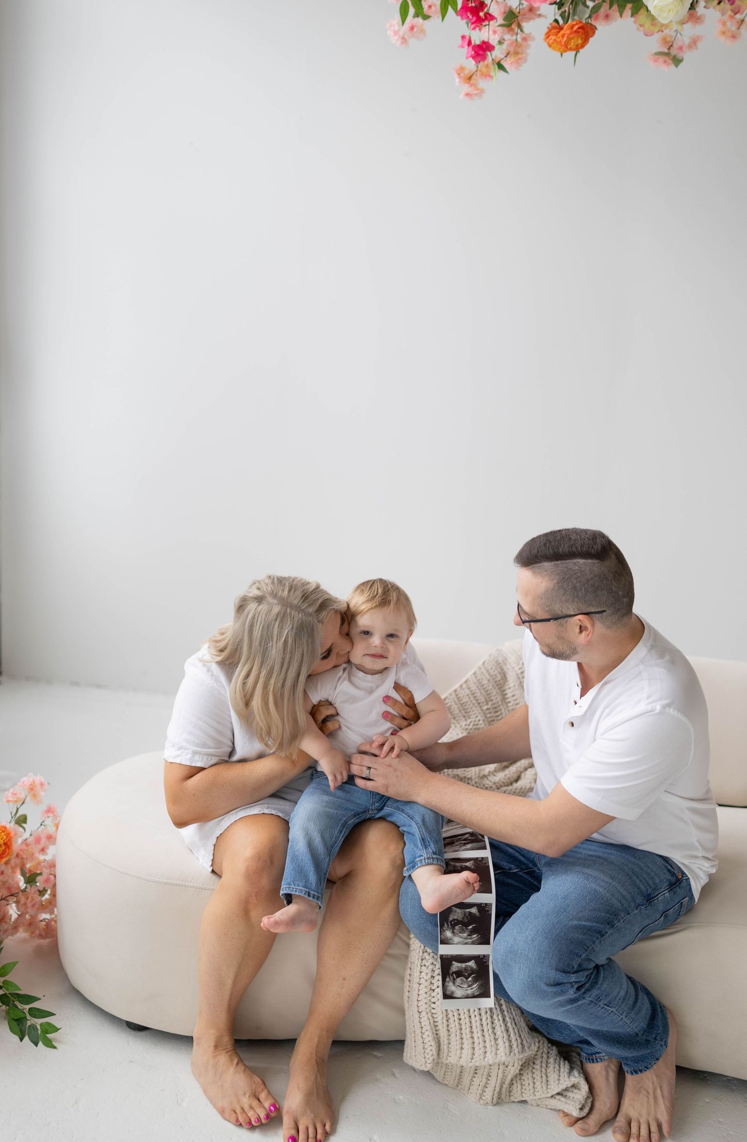 A family sits together on a white couch with ultrasound photos, celebrating a pregnancy announcement with floral decor above.