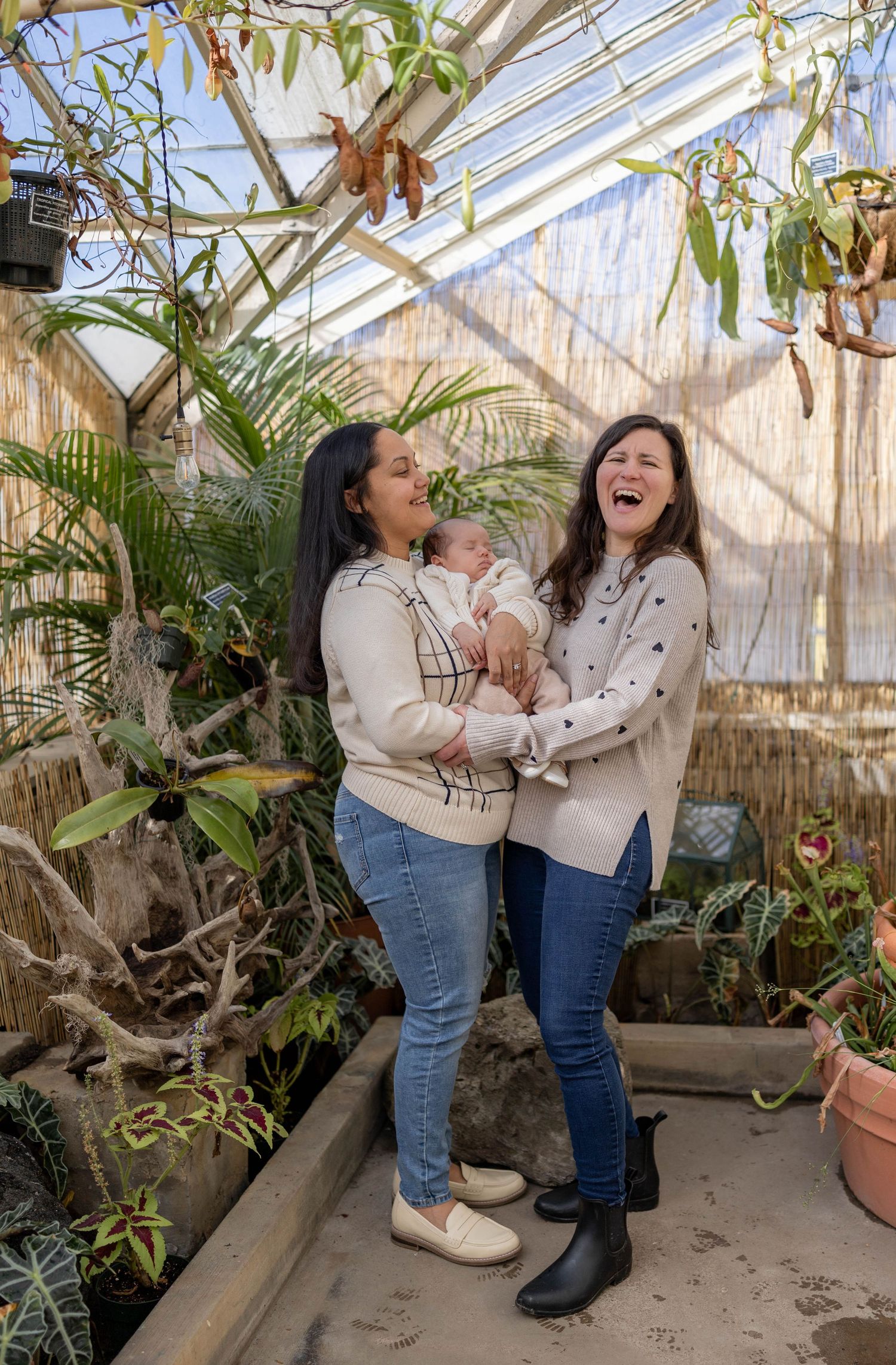 Two joyful friends sharing laughter together inside a sunlit greenhouse filled with tropical plants and greenery.