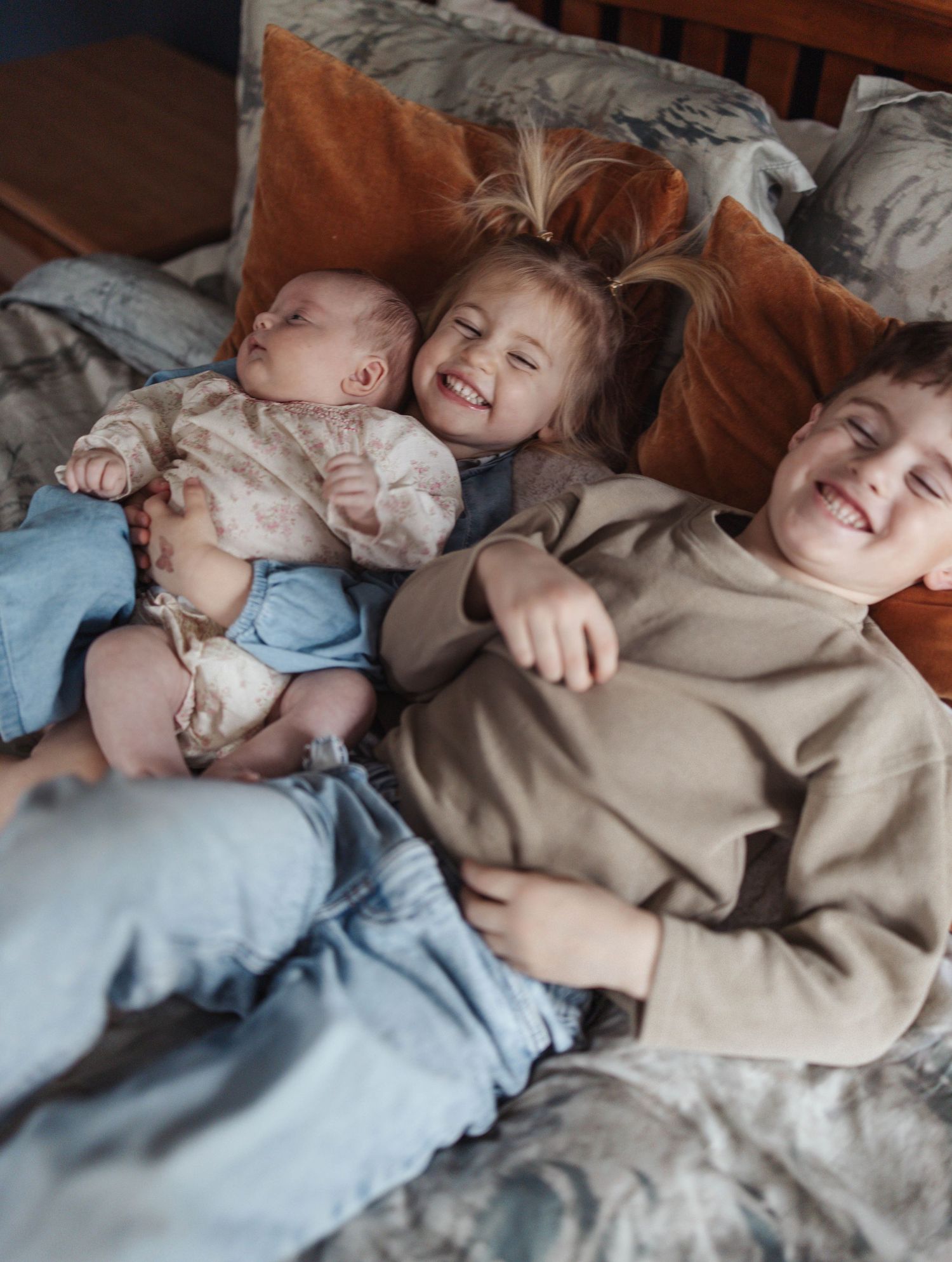 Three children relax and laugh together on a bed with cozy pillows and blankets.