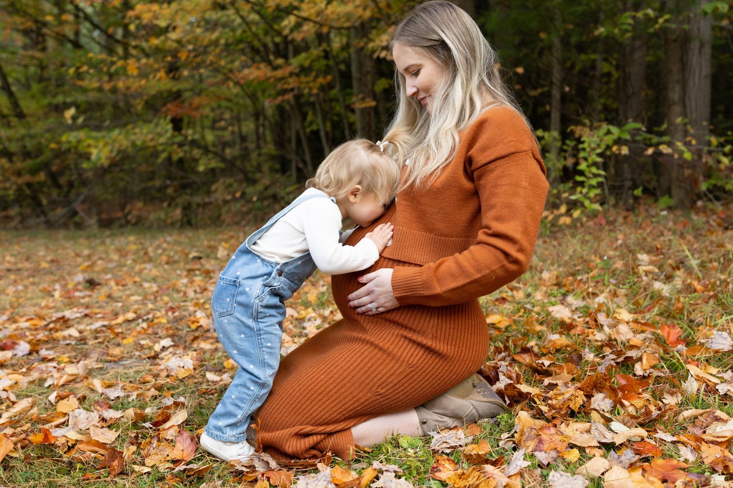 A parent in a rust-colored sweater dress kneels in autumn leaves with a small child in denim on a fall day.
