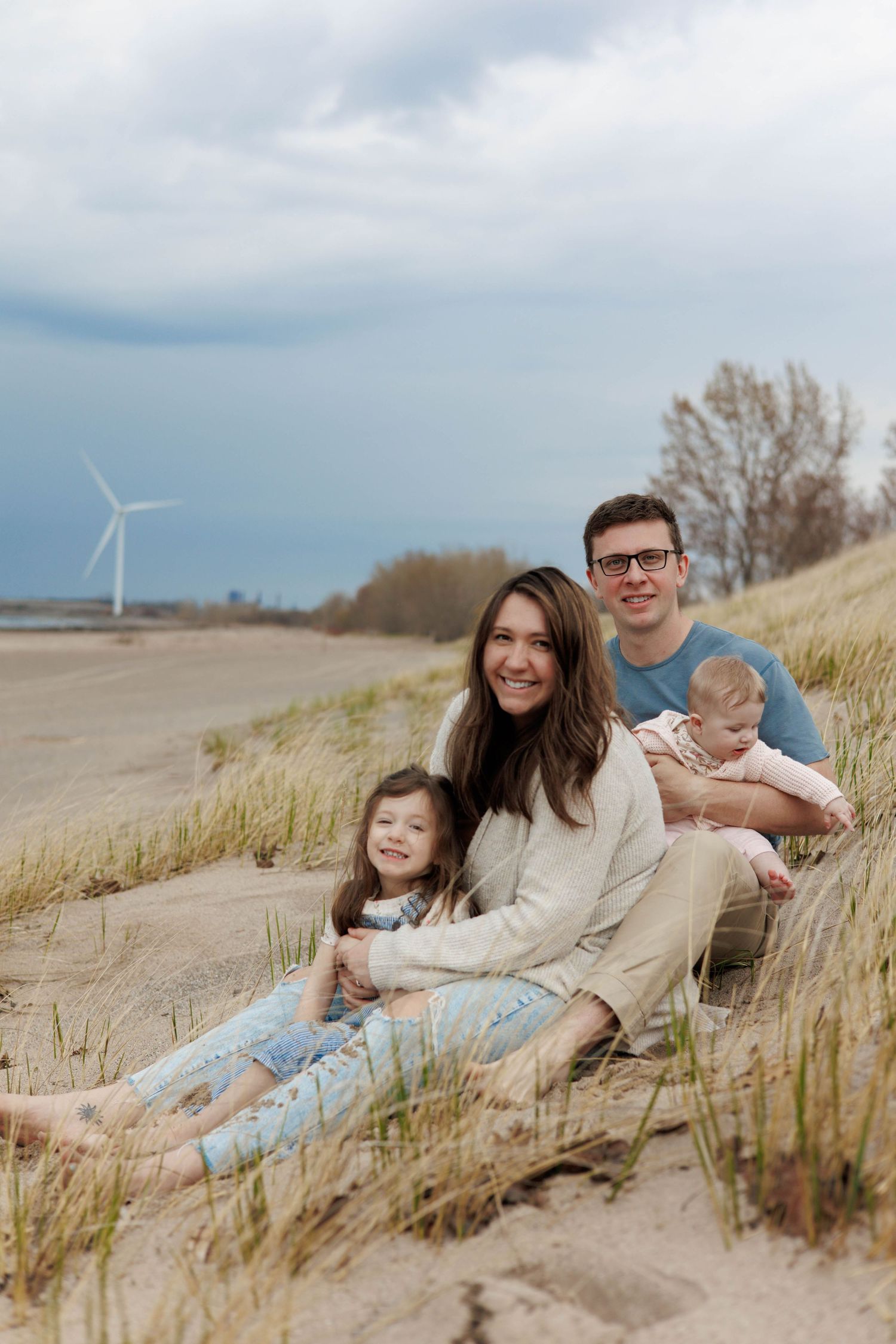 Family of four sitting on sandy beach dunes with a wind turbine in the background on a cloudy day.