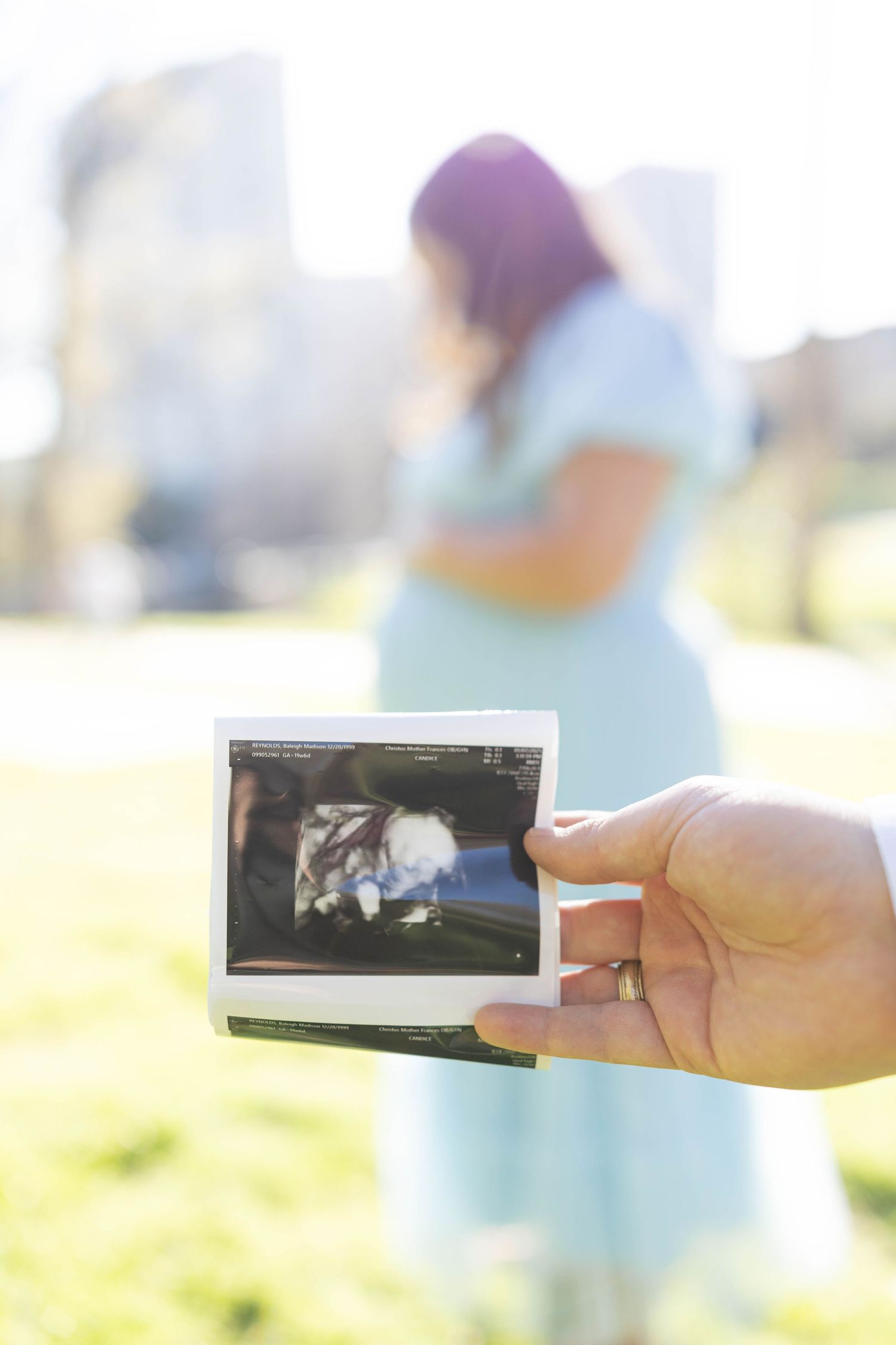A hand holds an ultrasound photo in the foreground while a pregnant person in a mint dress stands blurred in the background.