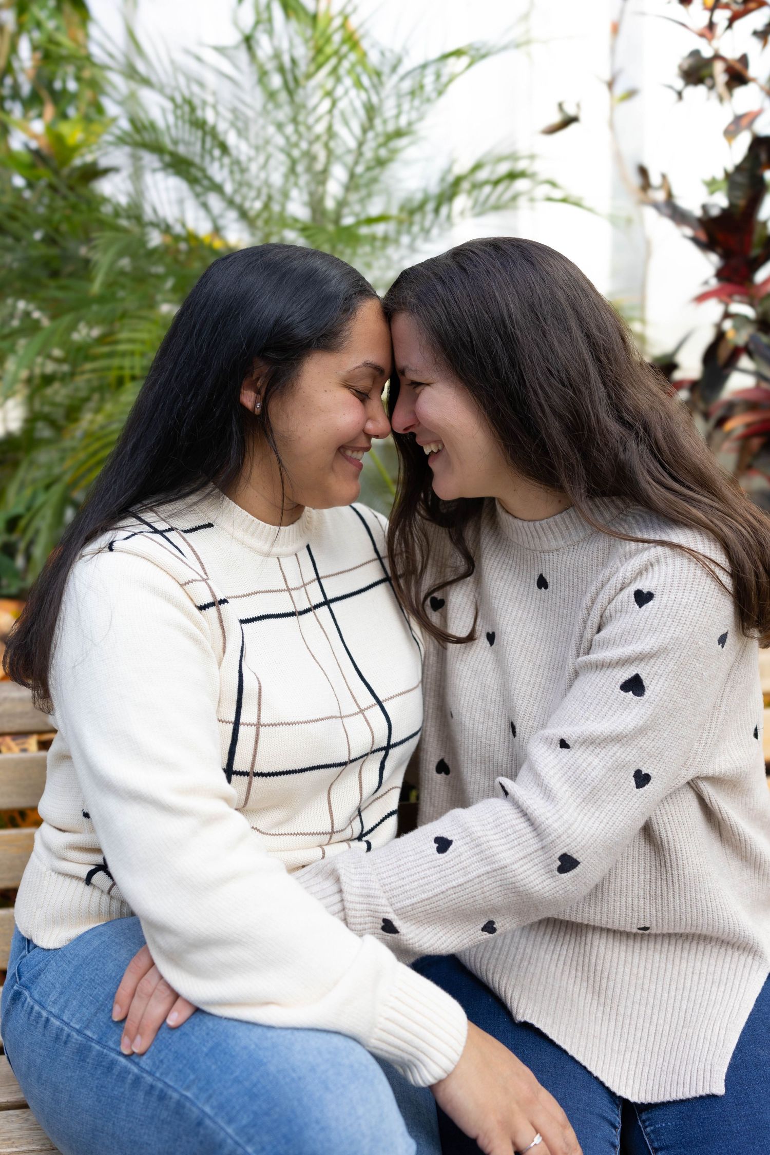 Two people in white sweaters share an intimate moment while sitting together against a tropical plant backdrop.